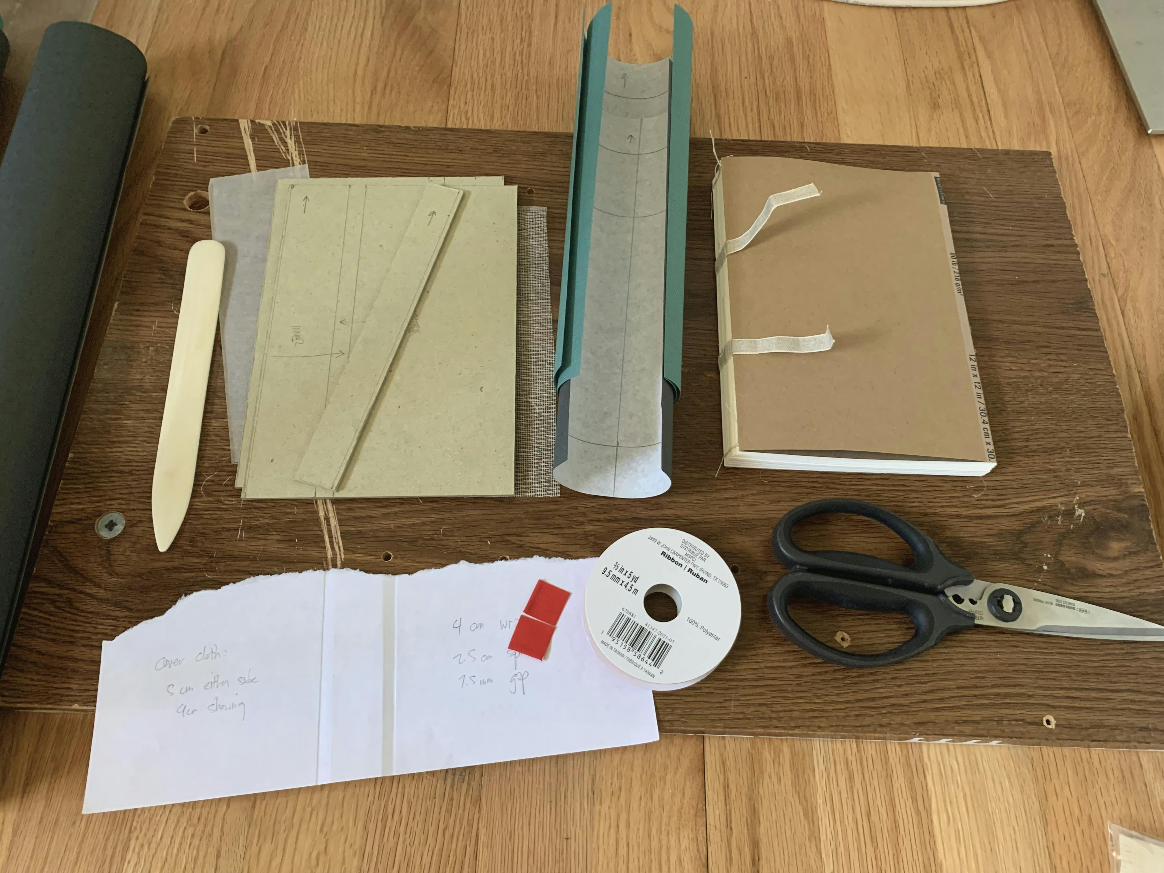 A photo of the parts of my notebook: cover boards, fabric, the sewn book block, scissors, ribbon, and a spine measuring guide rest on a wooden floor.