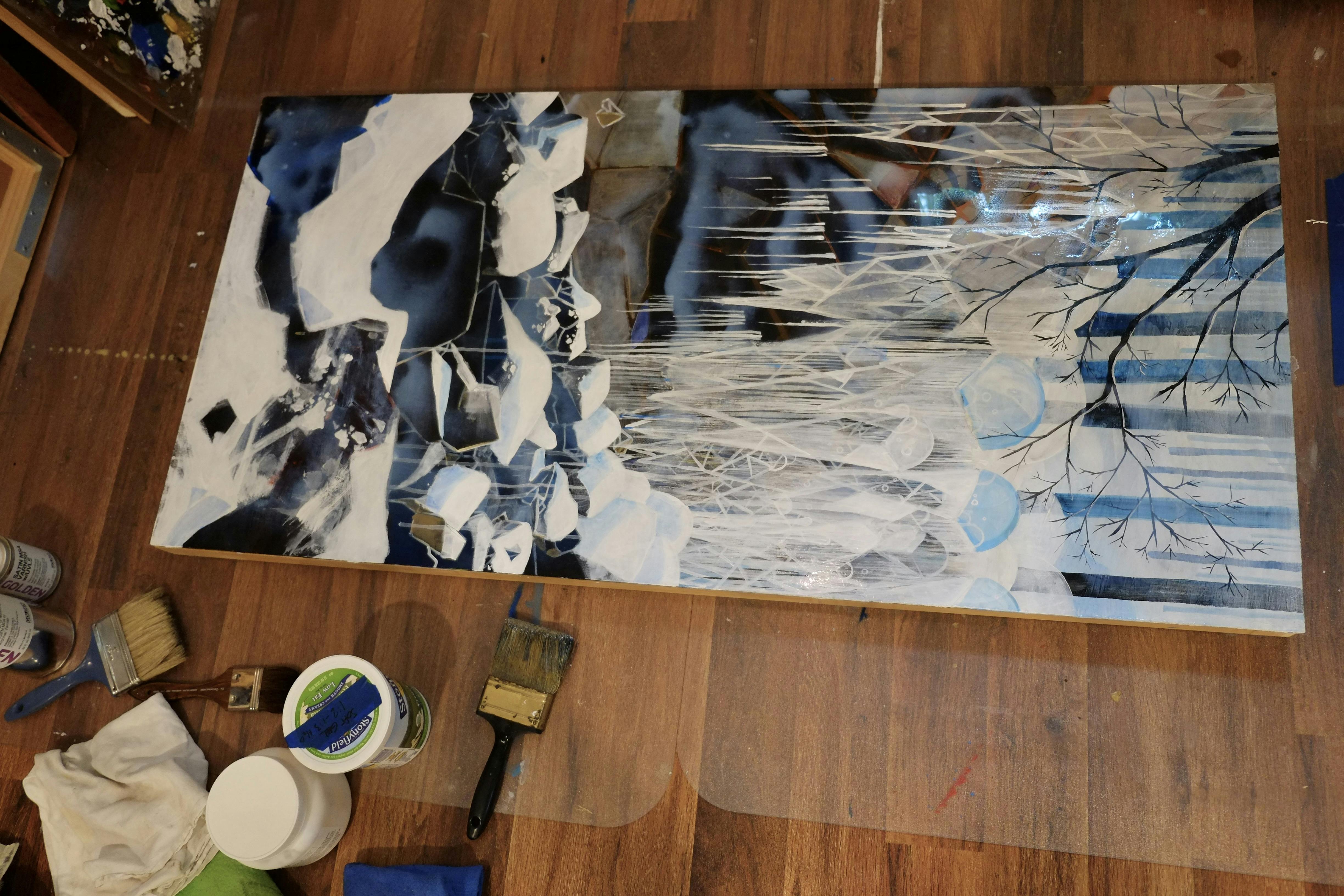 An overhead perspective of one of the panels of my Frozen Waterfall triptych. A wet coat of varnish has just been applied and is visible as cloudy puddles on the painting's surface. Brushes and yogurt tubs (containing varnish and solvent) are visible on the floor nearby.