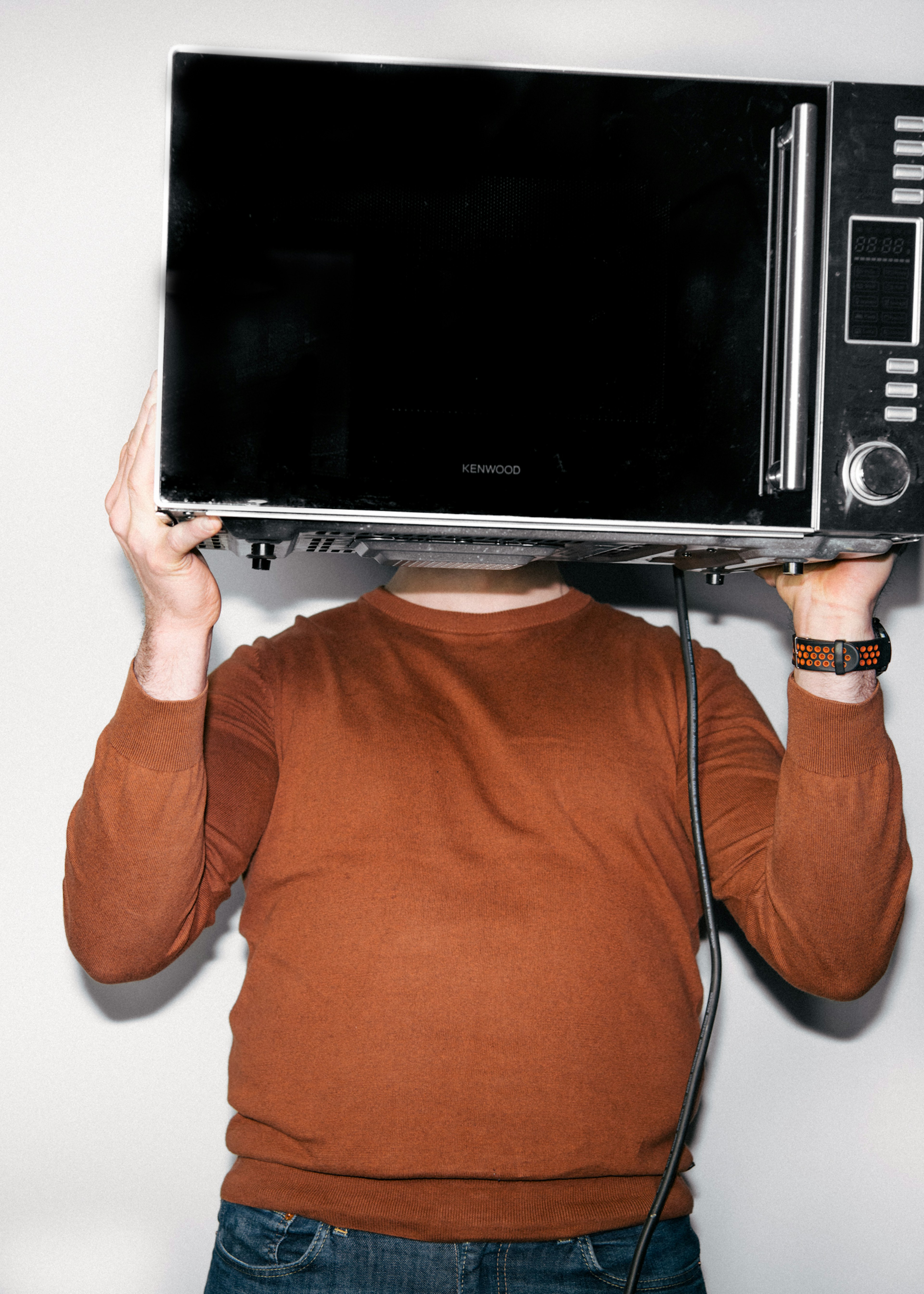 Einstaklingur í brúnum peysu heldur örbylgjuofni yfir höfuðið þannig að andlitið sést ekki.