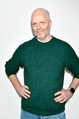 Sköllóttur maður með skegg í grænni peysu og snjallúri, stendur með hendurnar á mjöðmunum á hvítum bakgrunni og brosir af sjálfsöryggi.