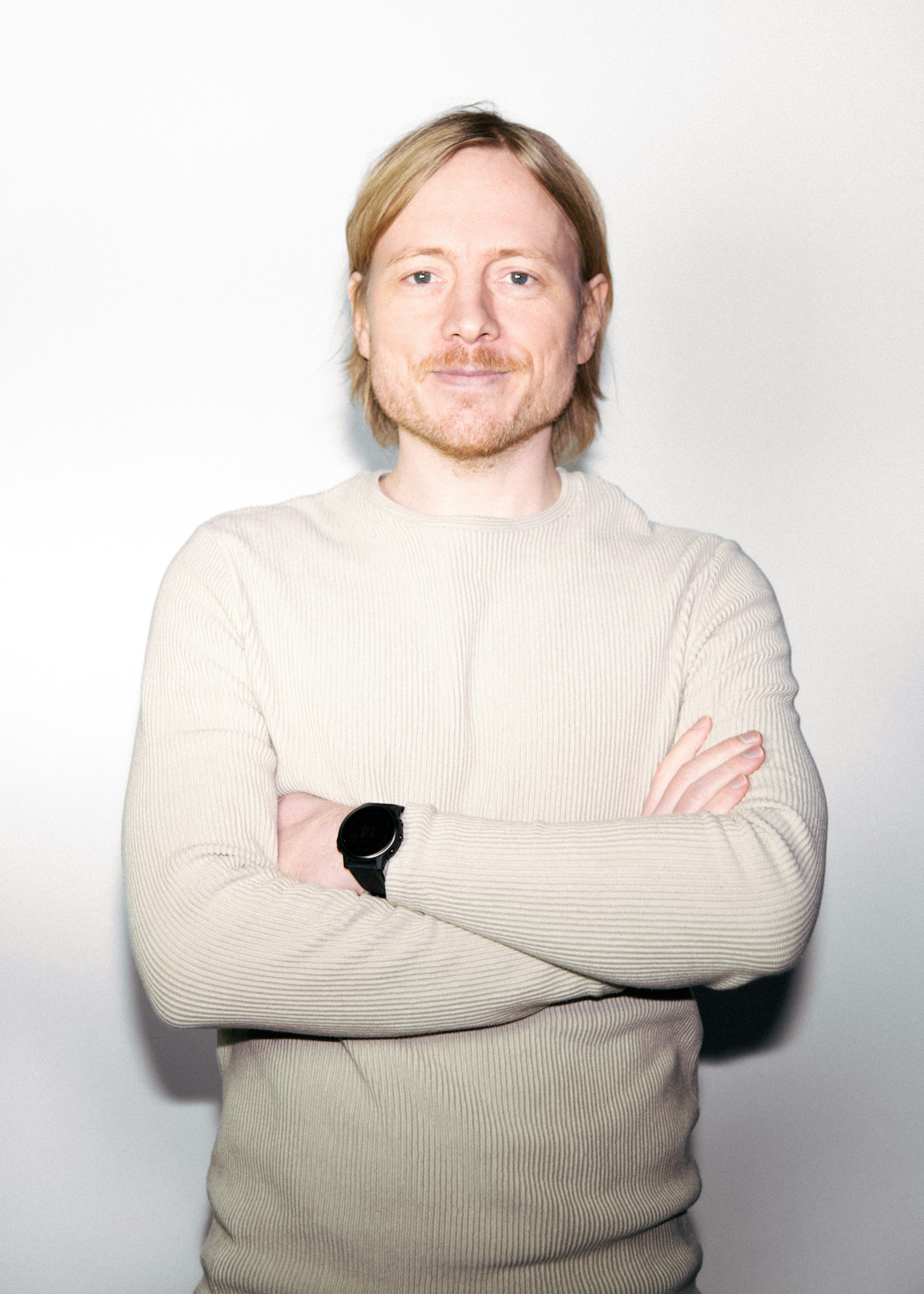 Maður með sítt ljóst hár stendur við hvítan bakgrunn, klæddur í létta peysu, með krosslagða hendur og sjálfstraustsfullt svipbrigði.