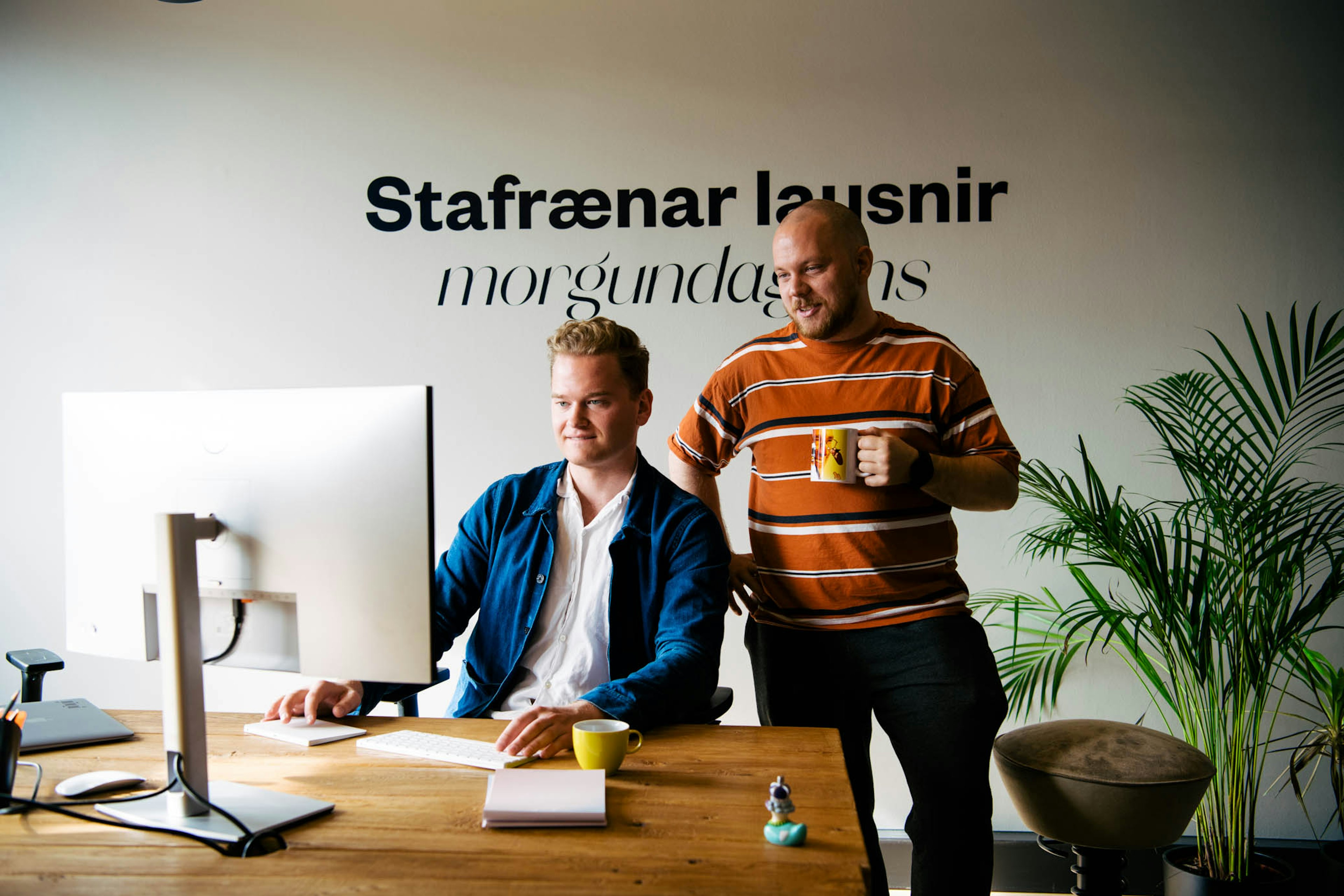 Tveir brosandi einstaklingar á skrifstofu, annar situr við tölvu og hinn stendur með kaffibolla.