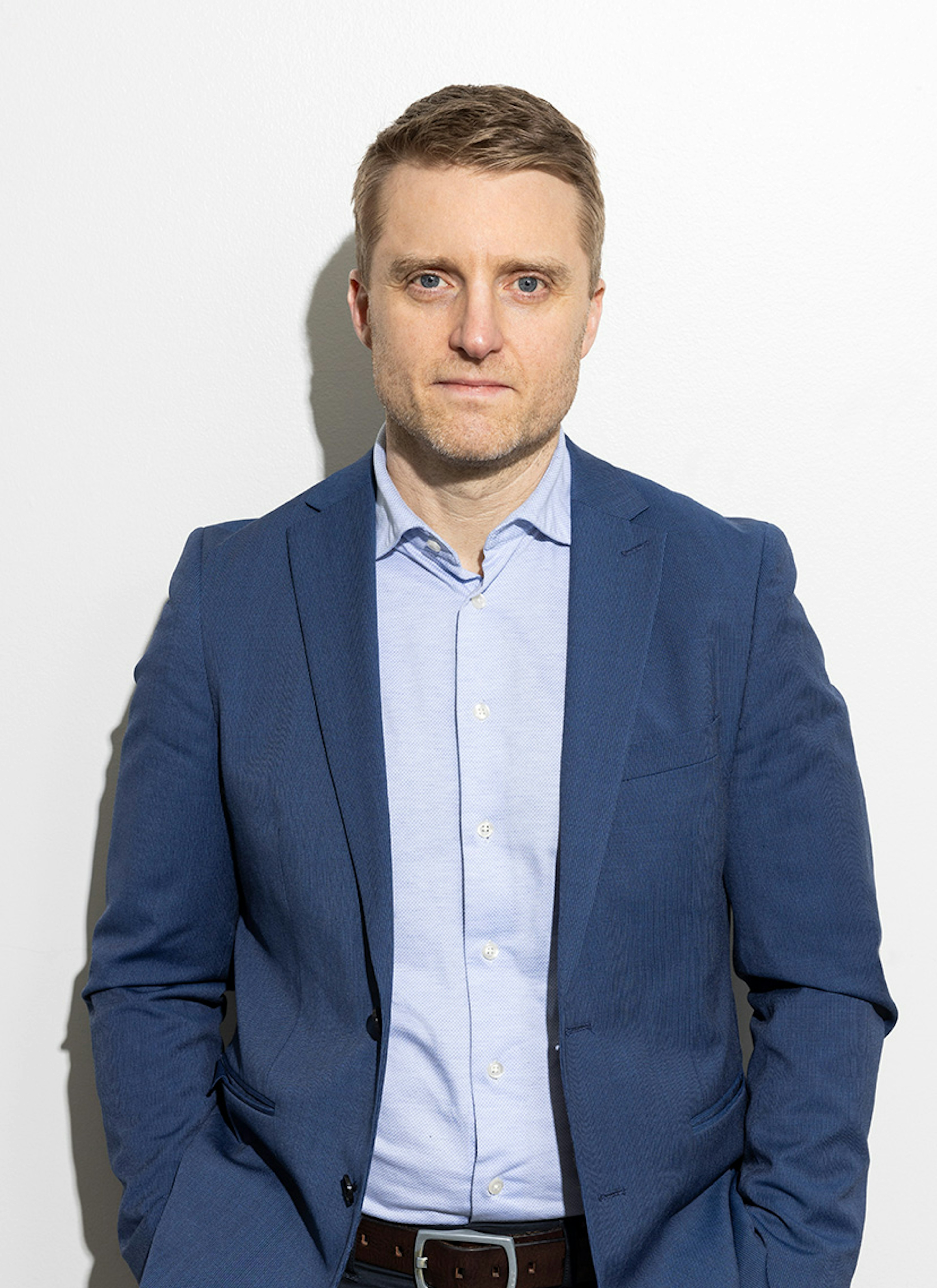 Maður í bláum jakkafötum og ljósbláum skyrtu situr fyrir á hvítum bakgrunni. Hann er með stutt hár og hlutlaust svipbrigði.