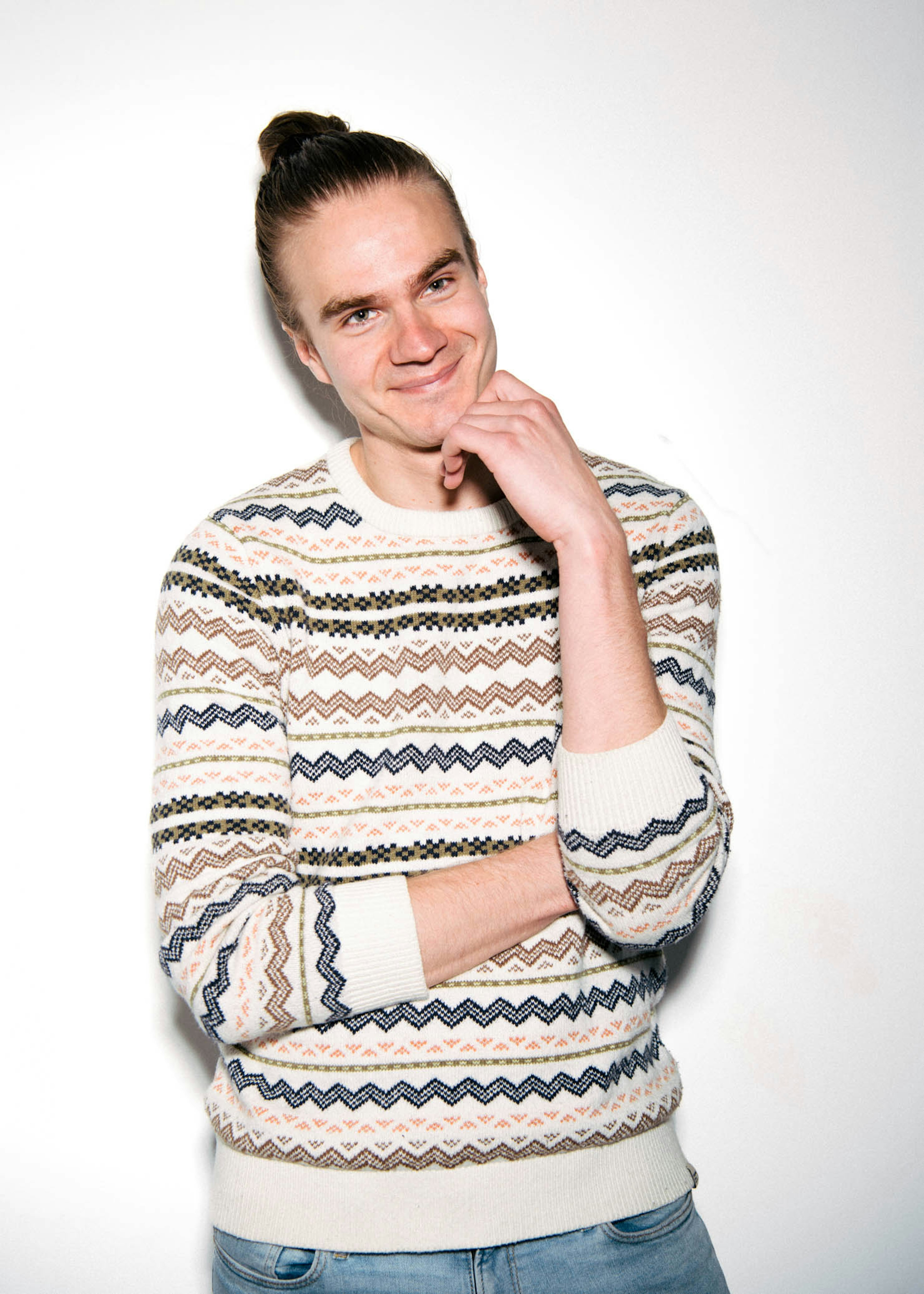 Einstaklingur með hugsandi svip, klæddur mynstraðri peysu og bláum gallabuxum, stendur fyrir framan einfalðan hvítan bakgrunn og geislar af afslöppuðu yfirbragði.