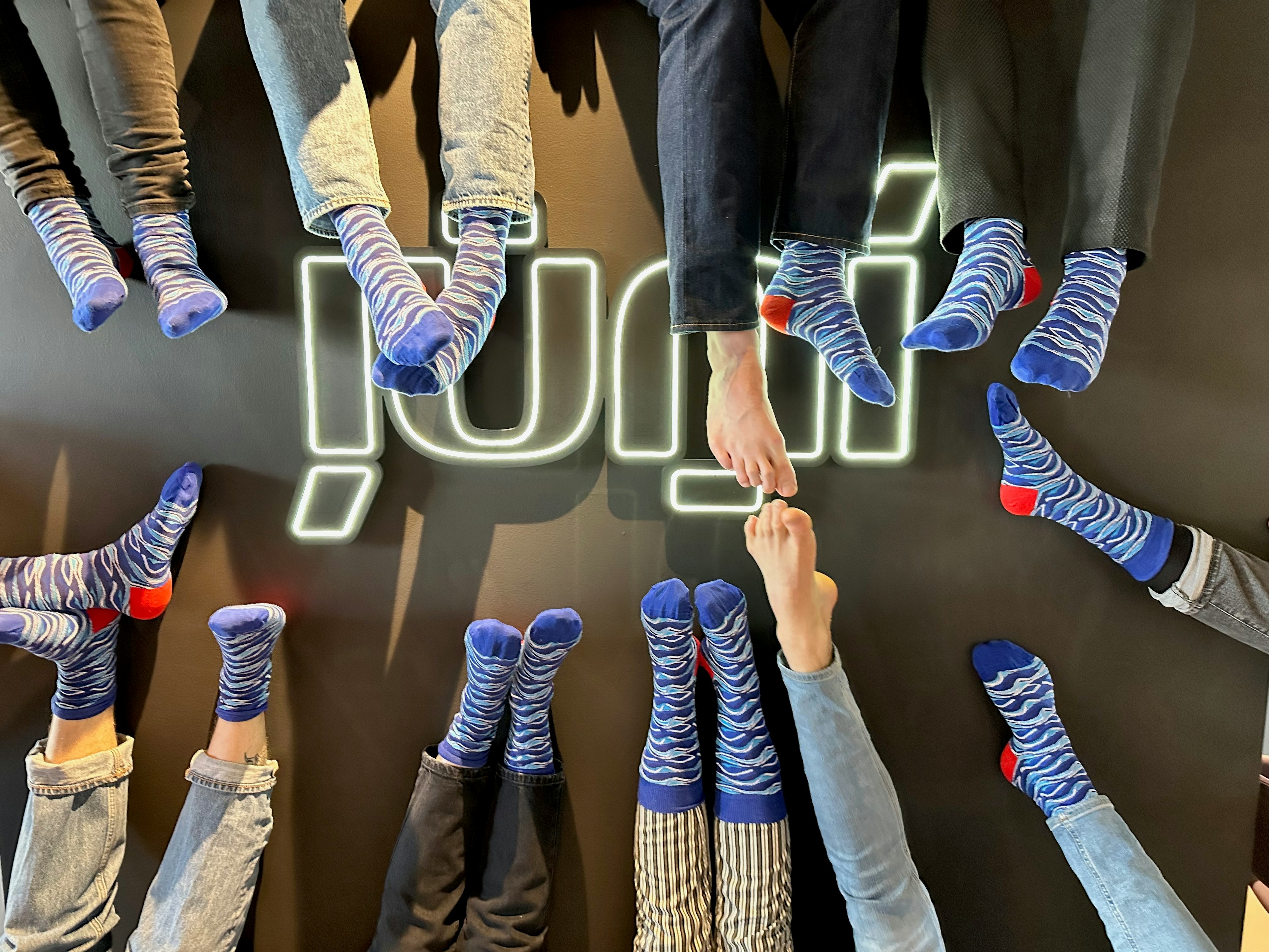 Fólk í litríkum bláum og rauðum sokkum liggur í hring með fætur vísandi inn á við gegn vegg með glóandi, stílfærðu Júní-merki í miðjunni.