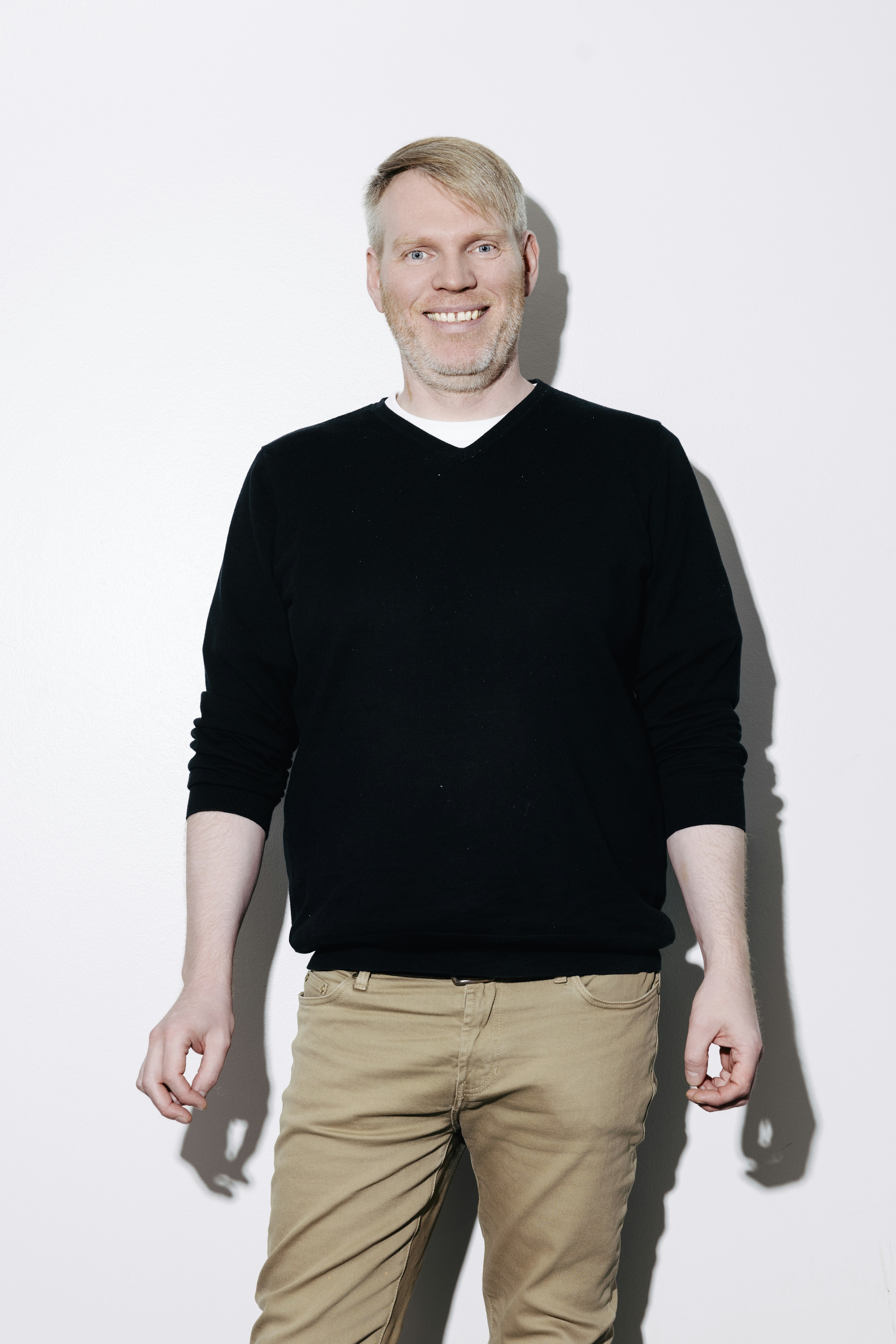 Maður stendur á hvítum bakgrunni, klæddur í svarta peysu og ljósbrúnar buxur.