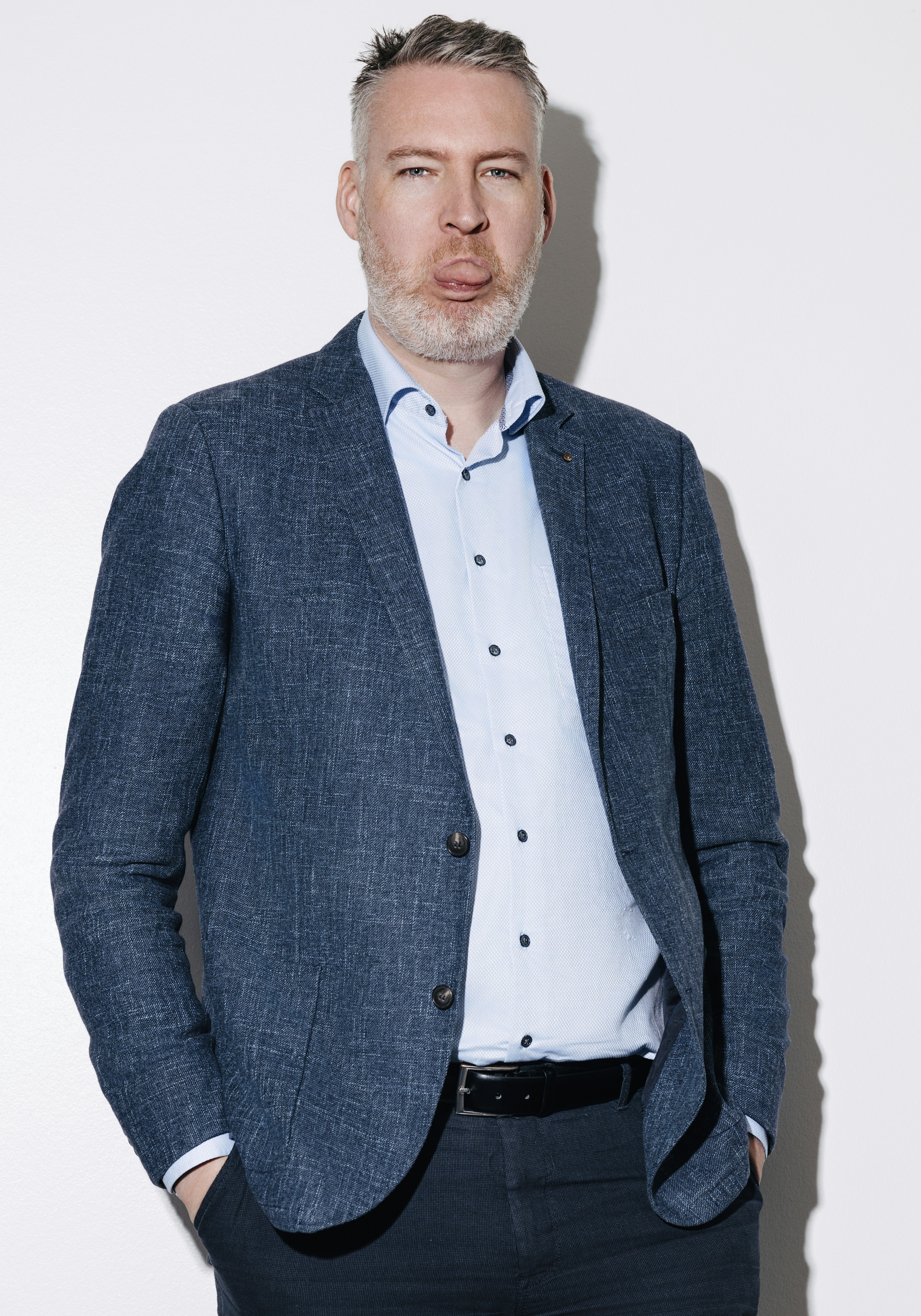 Maður með grátt skegg klæðist bláum jakka og ljósri skyrtu, stendur sjálfstraustsfullur með hendurnar í vösum.