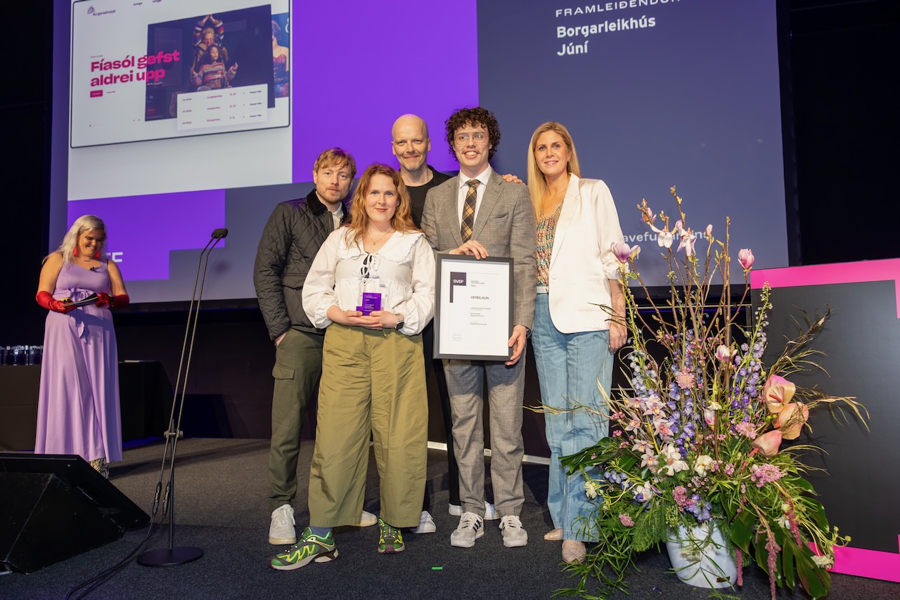 Júní-teymið stendur á sviði og brosir, heldur á ramma með SVEF-vottorði og SVEF-bikari, við hlið litríkrar blómaskreytingar, með stórri skjá og fjólubláu ljósi í bakgrunni.