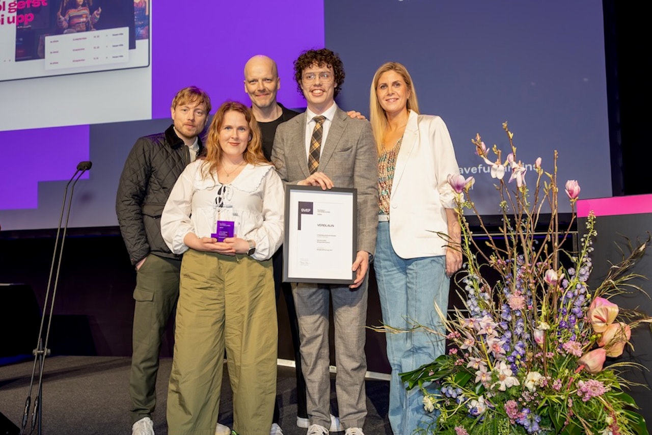 Júní-teymið stendur á sviði og brosir, heldur á ramma með SVEF-vottorði og SVEF-bikari, við hlið litríkrar blómaskreytingar, með stórri skjá og fjólubláu ljósi í bakgrunni.