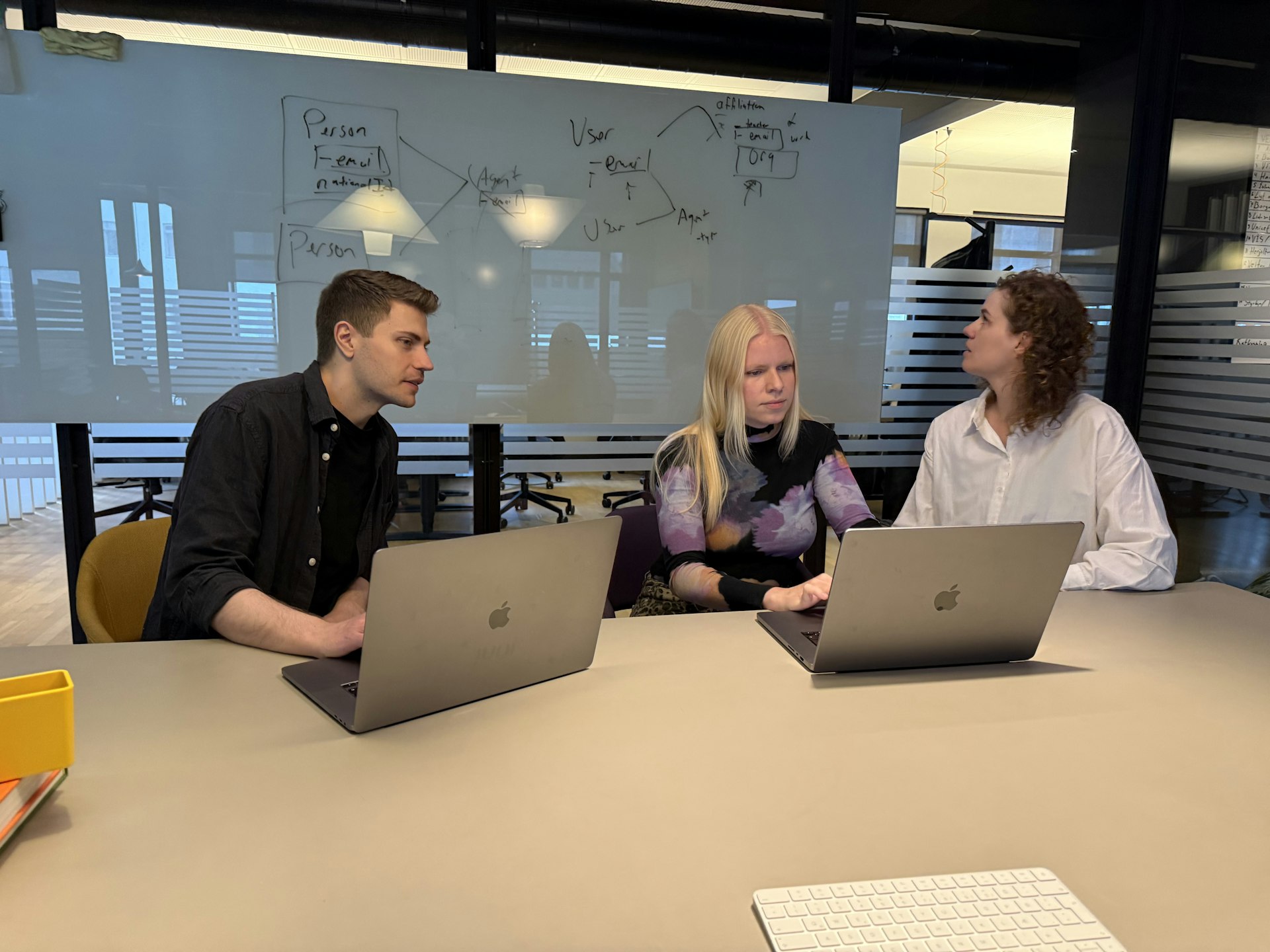 Þrír samstarfsaðilar sitja við borð með fartölvur og ræða verkefni, með teikningu á töflu í bakgrunni