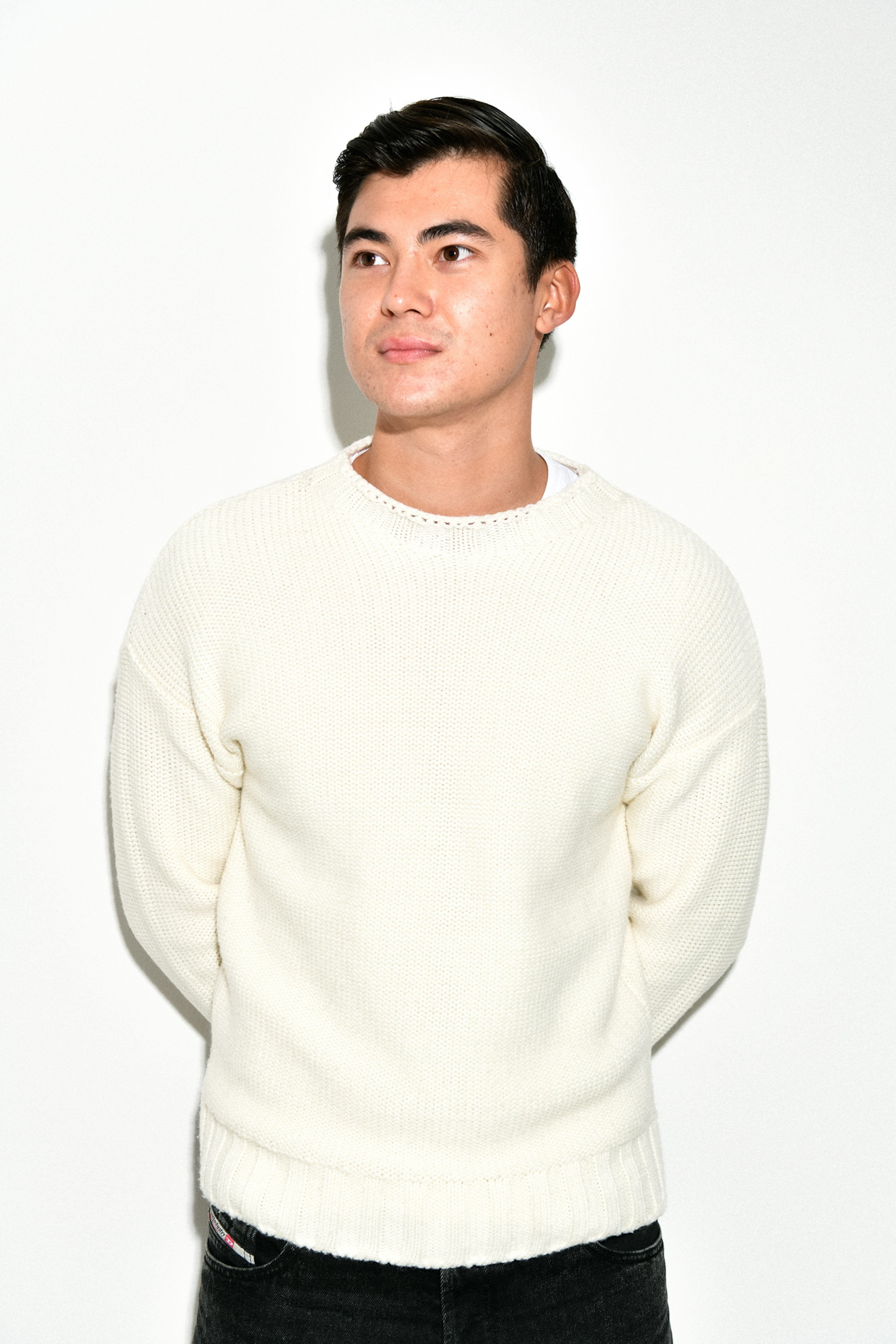 Einstaklingur í hvítum peysu stendur fyrir framan einfalðan hvítan bakgrunn og lítur til hliðar með hugsandi svip.