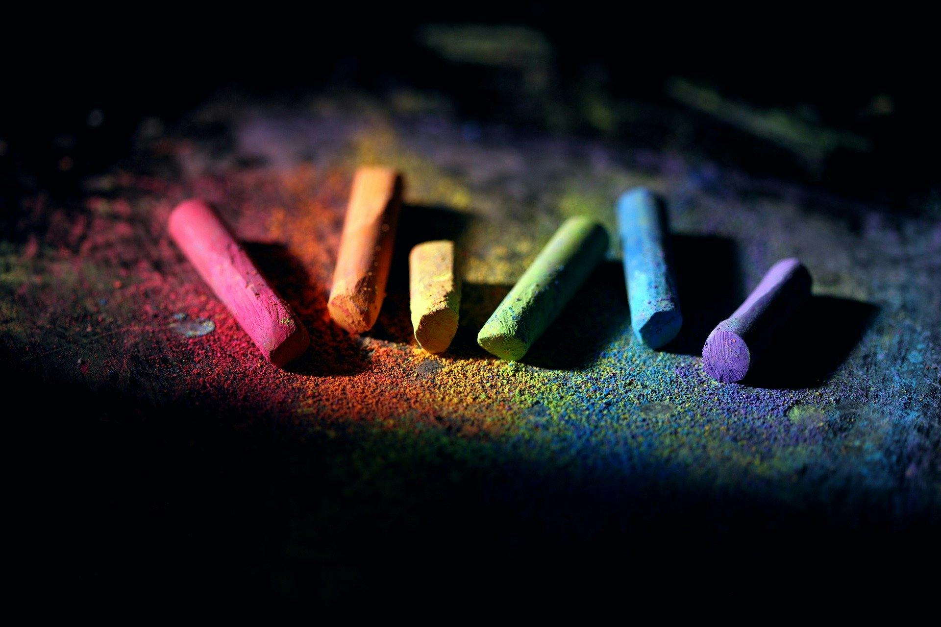 Six sticks of chalk laying on the ground, red, orange, yellow, green blue and purple