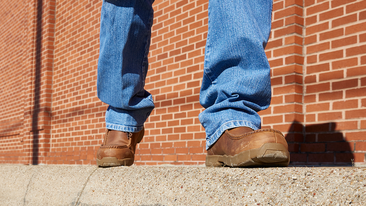 Un hombre con jeans camina frente a una pared de ladrillos rojos usando las Justin Light Duty Casuals.
