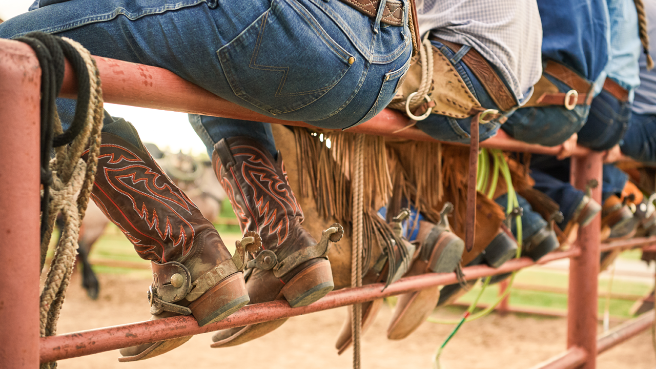 Multiple cowboys sitting on top of a fence with their boots resting on a lower rail.