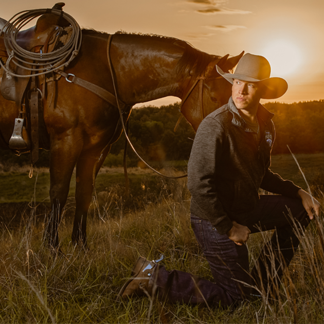 Professional Bull Fighter Cody Webster kneeling in a field while leading his horse on his ranch at sunset.