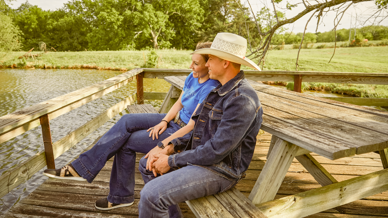 Professional Bullfighter Cody Webster and his wife Ashley sitting on a picnic table overlooking a pond at their ranch.