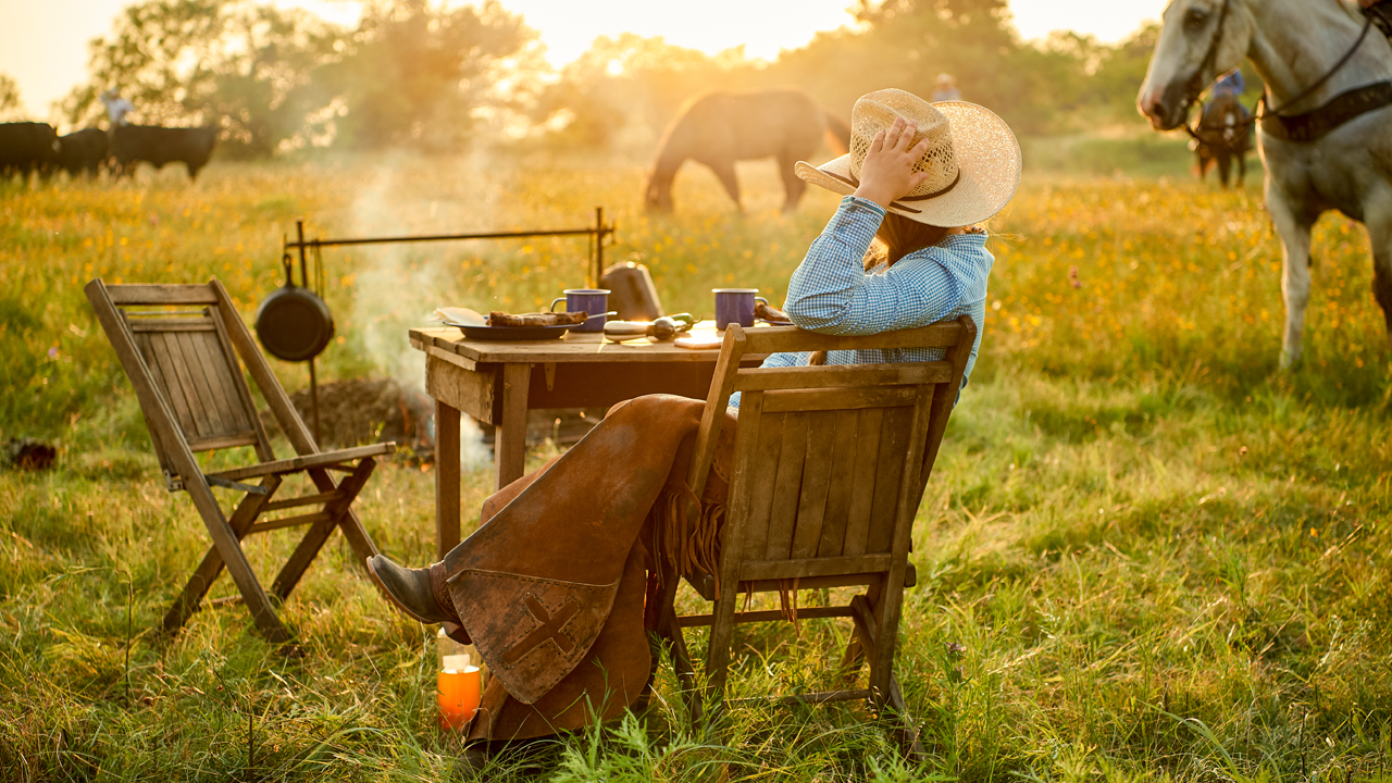 A lone cowgirl sitting at a table in the middle of a field overlooking a campfire at sundown.