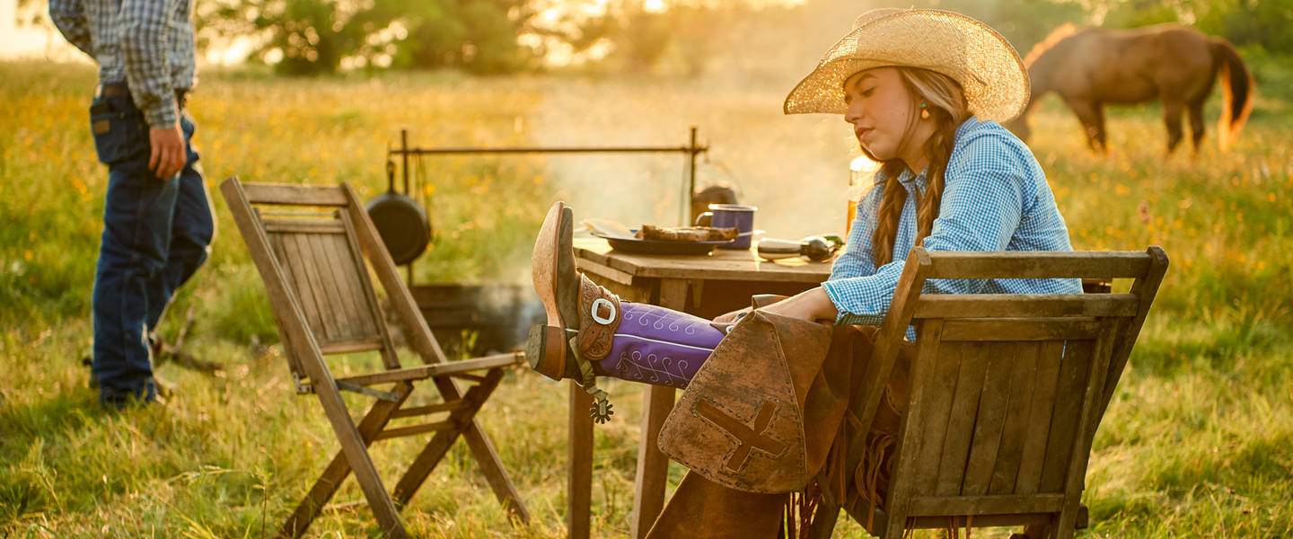 A lone cowgirl inspecting her brightly colored boot top while sitting at a table by a campfire, in an open field while a horse grazes in the background.