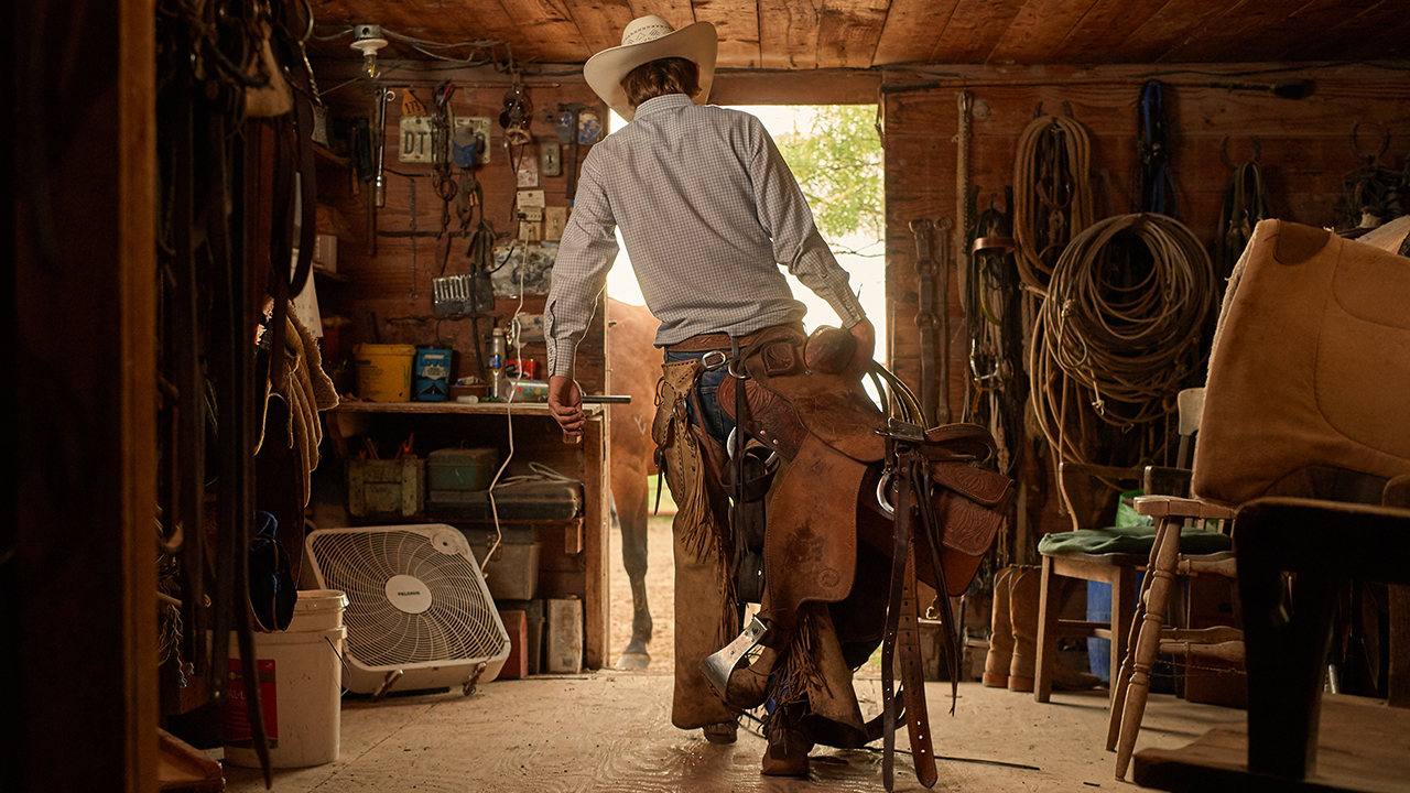 A cowboy walking away from the camera carrying a saddle from his tack room.