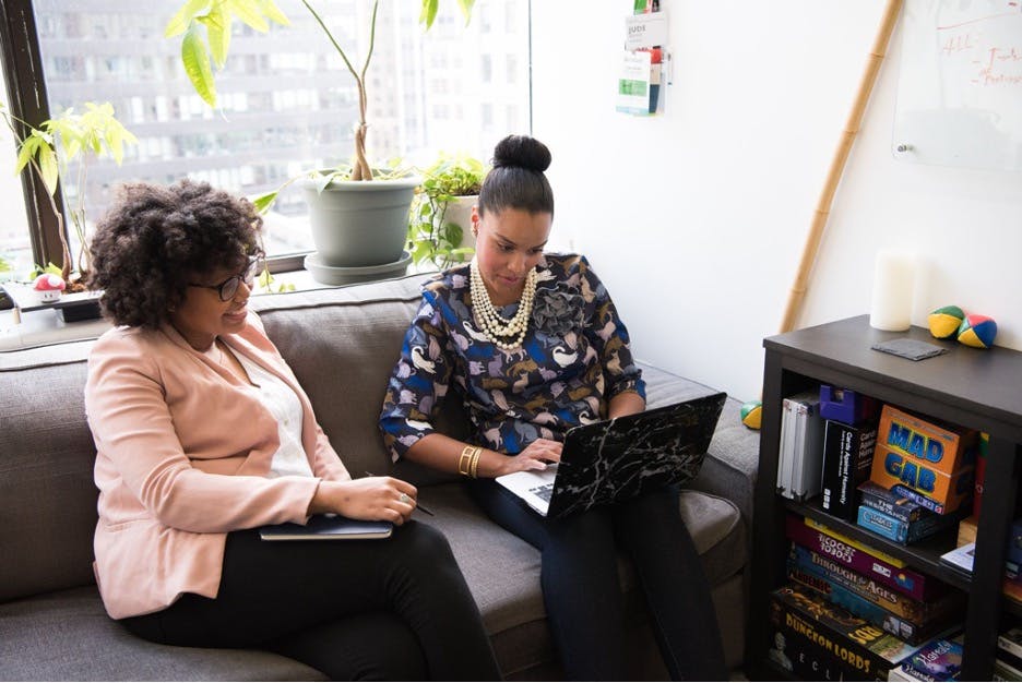 Woman with a laptop speaking to another woman holding a notebook