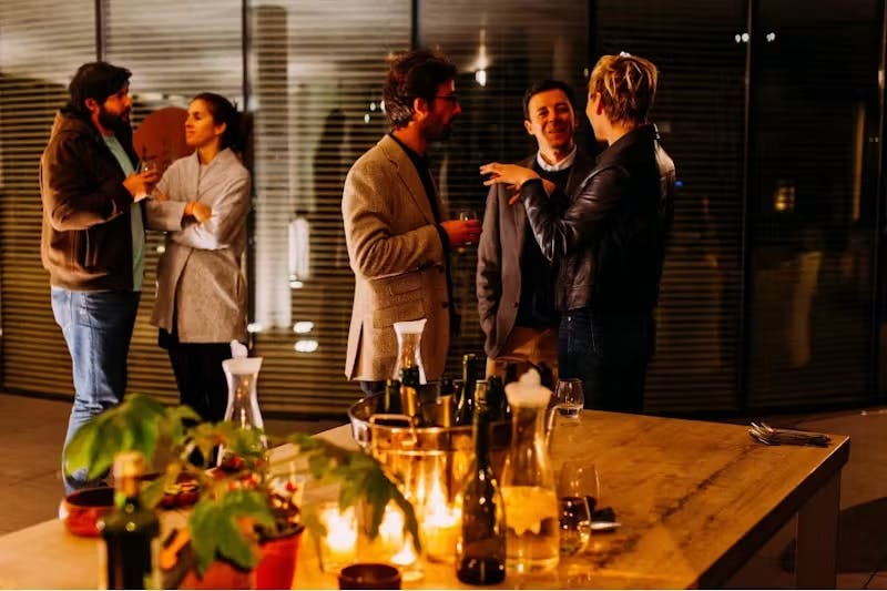 5 people chatting in two groups next to a table with wine bottles