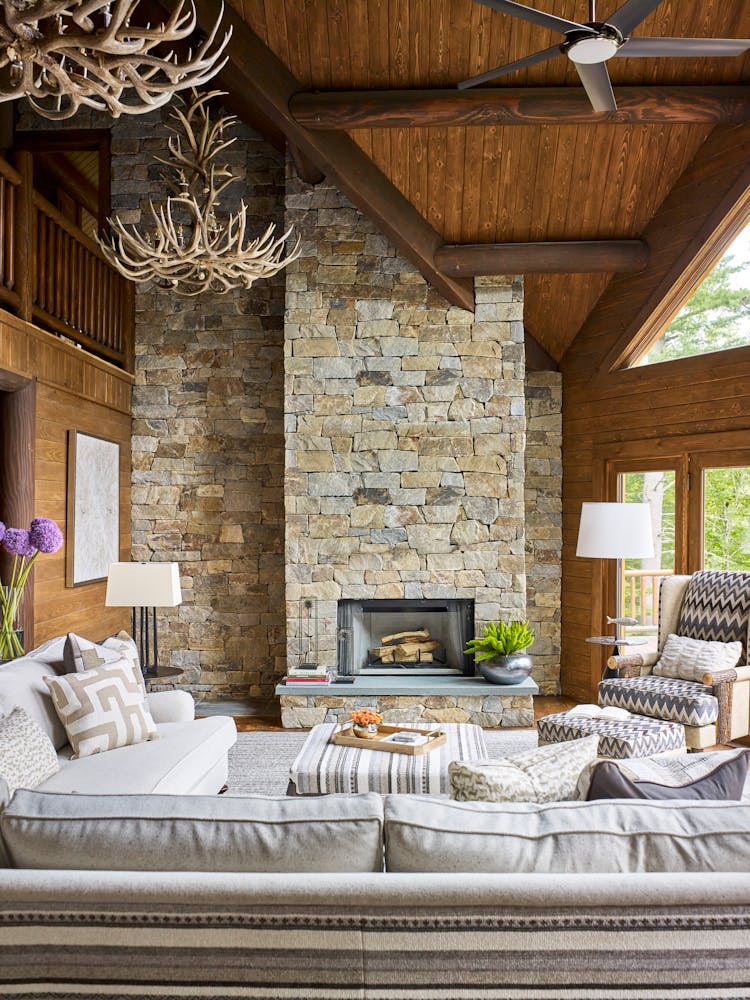 Spacious family room in a Hudson Valley log cabin featuring a double-height stone fireplace, vaulted wood ceilings, and expansive windows, designed by K.A. Murphy Interiors.