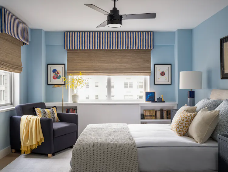 Guest bedroom with soft blue walls, layered bedding, custom window treatments, and a reading chair by K.A. Murphy Interiors.