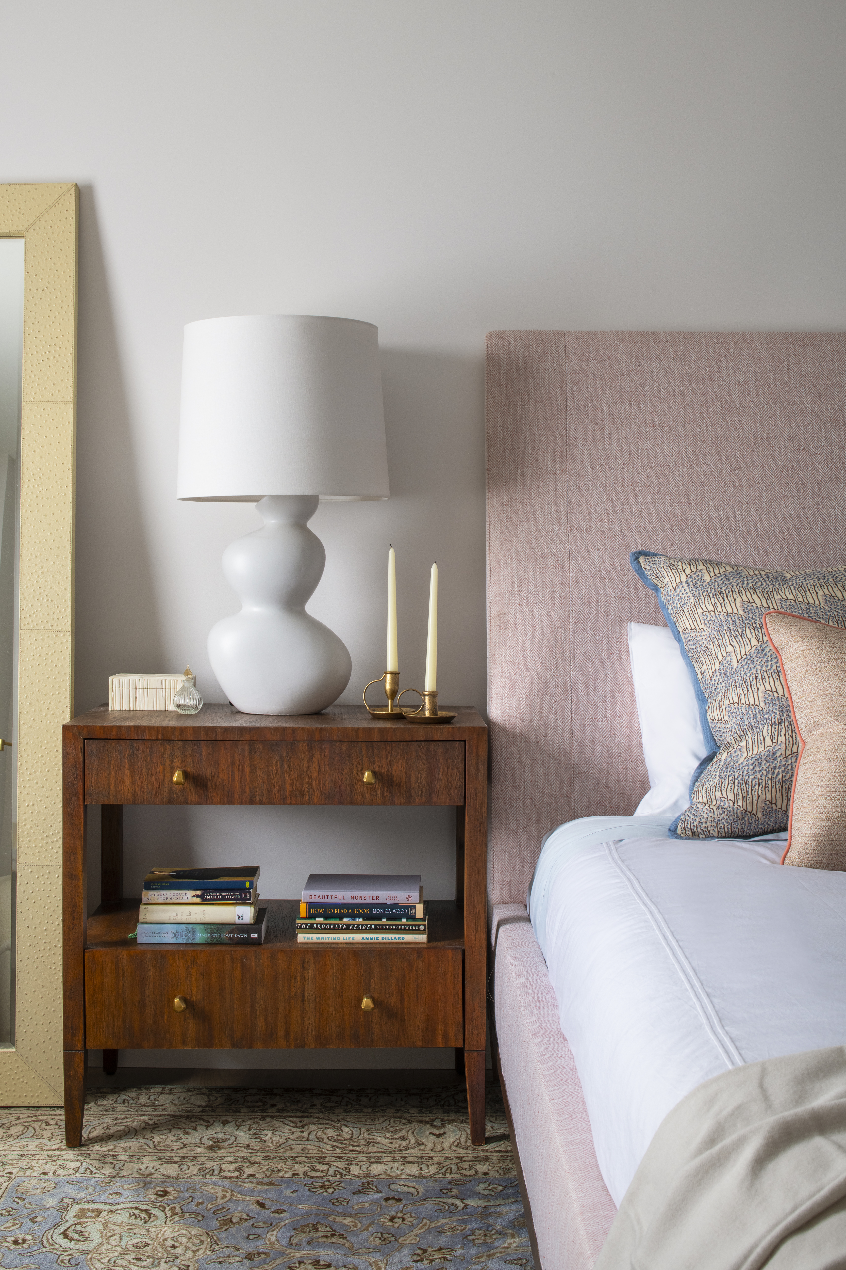 Primary bedroom nightstand with sculptural lamp, stacked books, and soft pink upholstered headboard by K.A. Murphy Interiors.