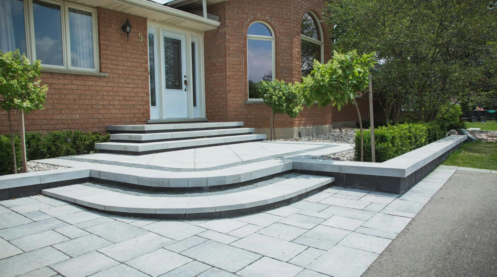 Gorgeous grey front porch and patio area built with interlocking stone. Leading edge of lower two steps consist of an oval corner configuration leading to an open porch area. Additional rectangular three steps lead up to the front door of the home. 