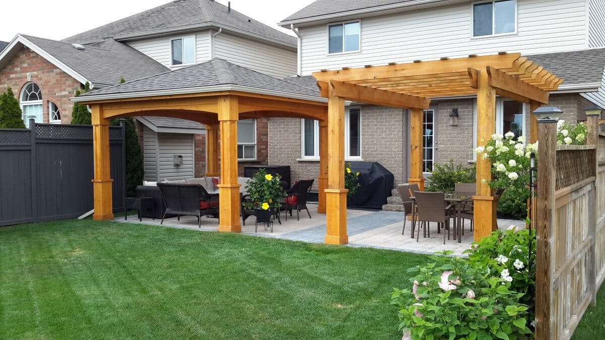 Interlocked grey and sand colour stone patio. Lush, green lawn meets patio edge. Two wooden pergolas rest side by side on the stone patio. One pergola provides full shade with an enclosed roof, while the other consists of wood beam roof - spaced to allow some sunlight through.   
