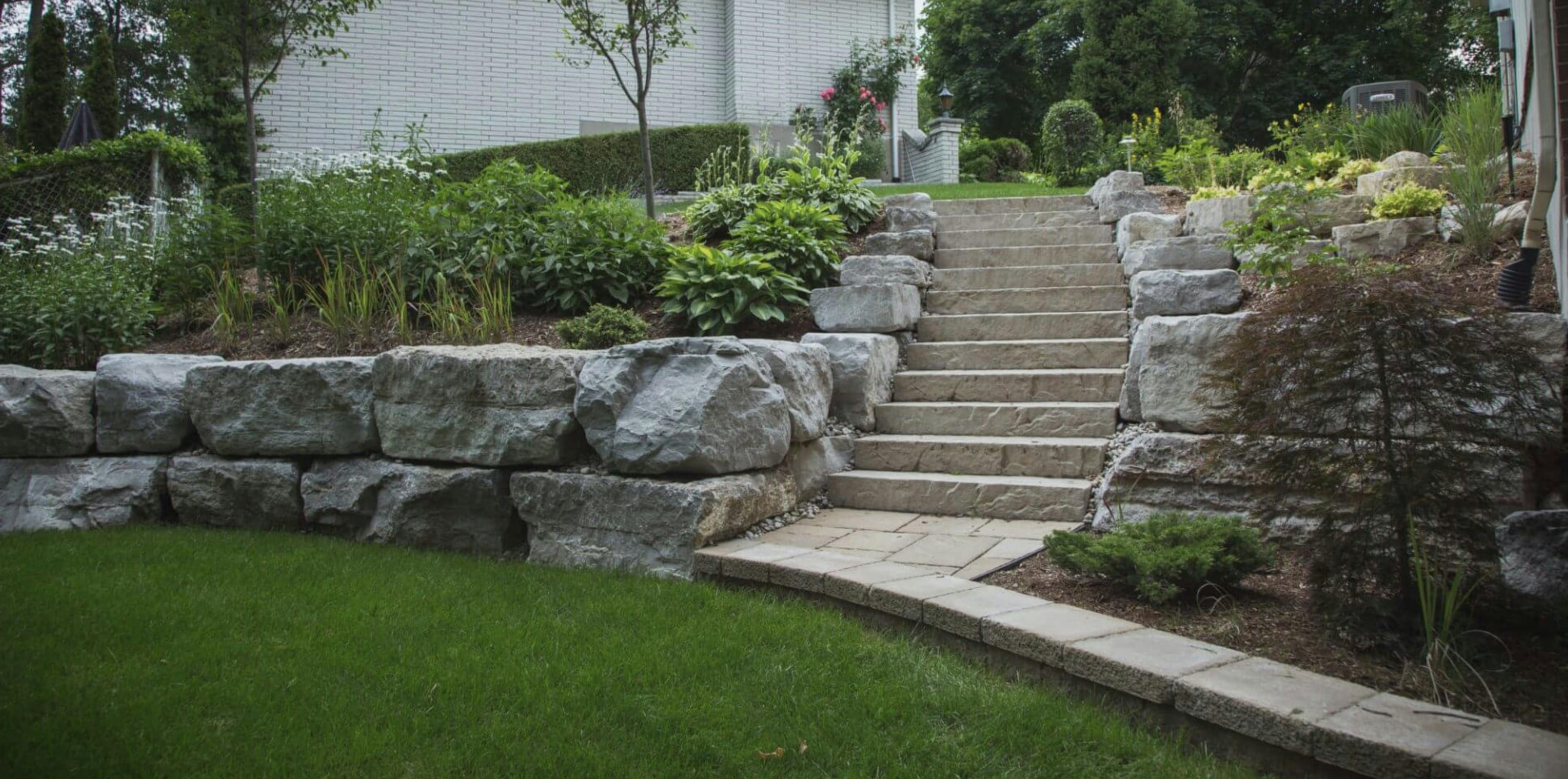 Twelve natural stone stairway leads up from lower backyard area to upper backyard area. Large natural stone blocks flank the stairway on both sides and provide a retaining wall at the edge of the lower yard. Garden containing a variety of plants flank both sides of the staircase. 