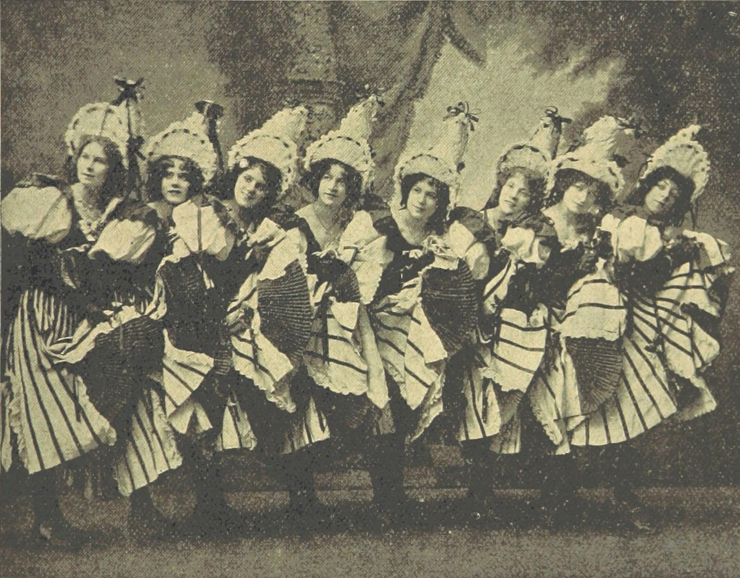 A black-and-white, old-fashioned photo of a chorus line of women wearing full, striped skirts and tall, ribboned hats. 