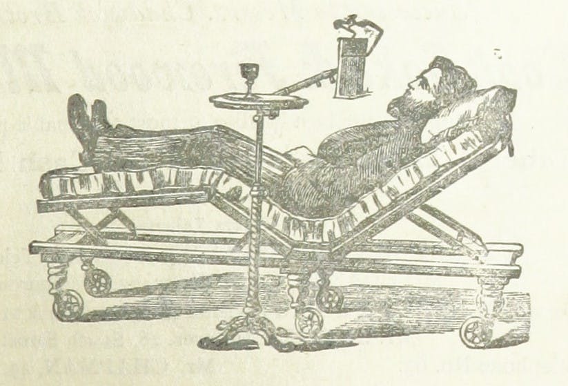 A man in Edwardian clothing and a beard reclines in a medical chair, with a small table with an instrument attached to it by his side.