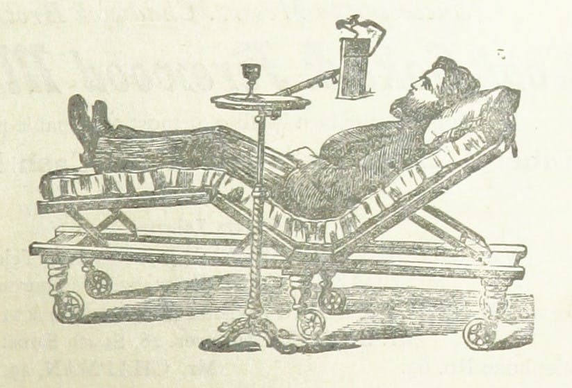 A man in Edwardian clothing and a beard reclines in a medical chair, with a small table with an instrument attached to it by his side. 