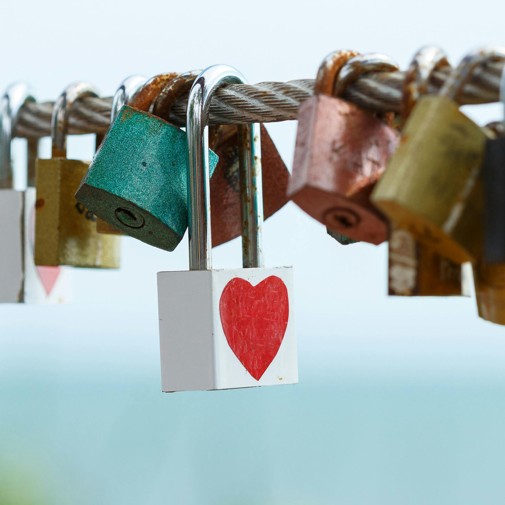 Colourful padlocks secured to a rope. A prominent white padlock has a red love heart drawn onto it.