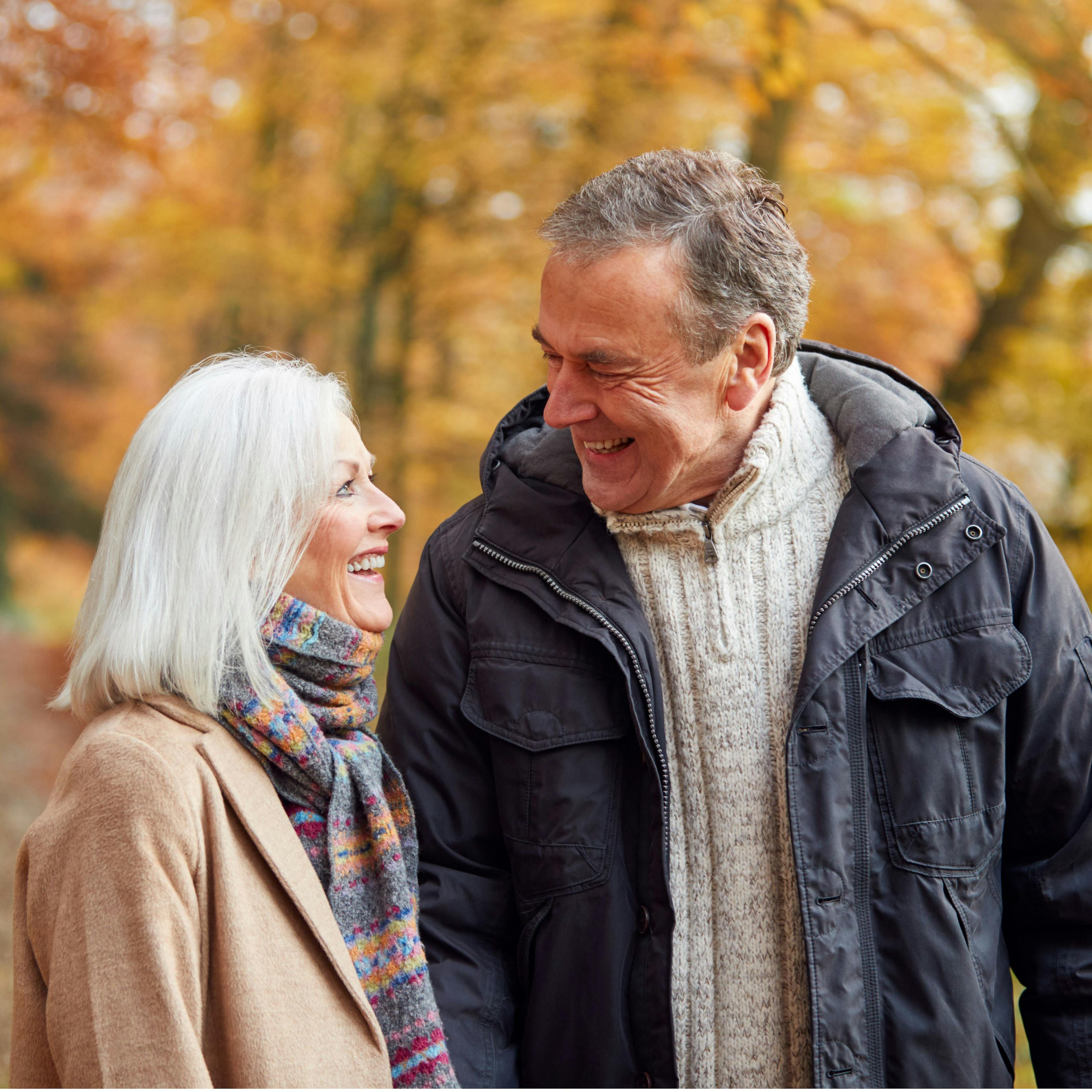 Grey-haired couple smile at each other on autumn walk.