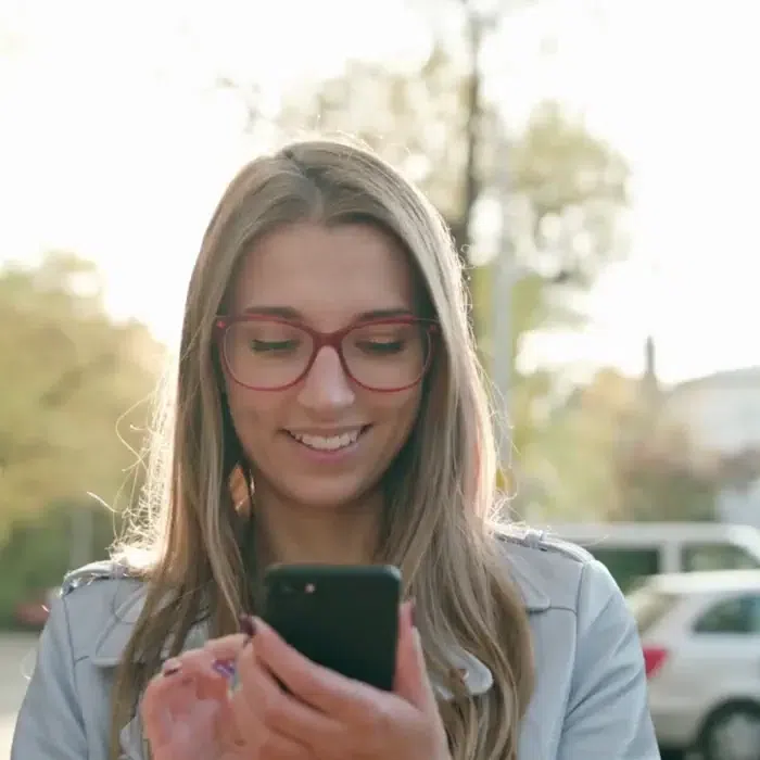 Girl in glasses smiles while checking her messages as the sun shines behind her