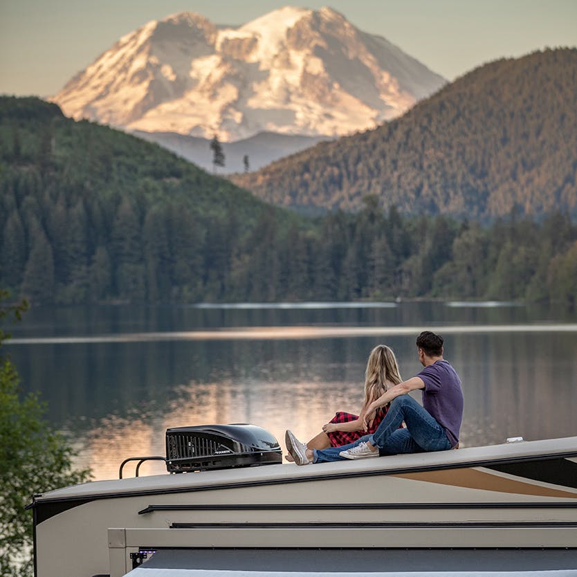 Couple sits atop a fifth wheel looking out at a mountain and lake