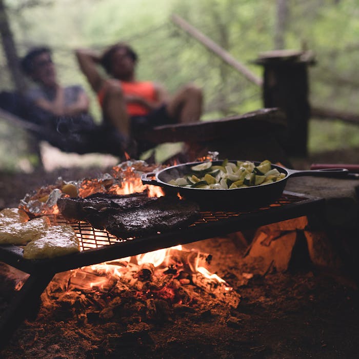 Couple relaxing on a hammock in front of campfire with a grate cooking steaks and vegetables in cast iron skillet 