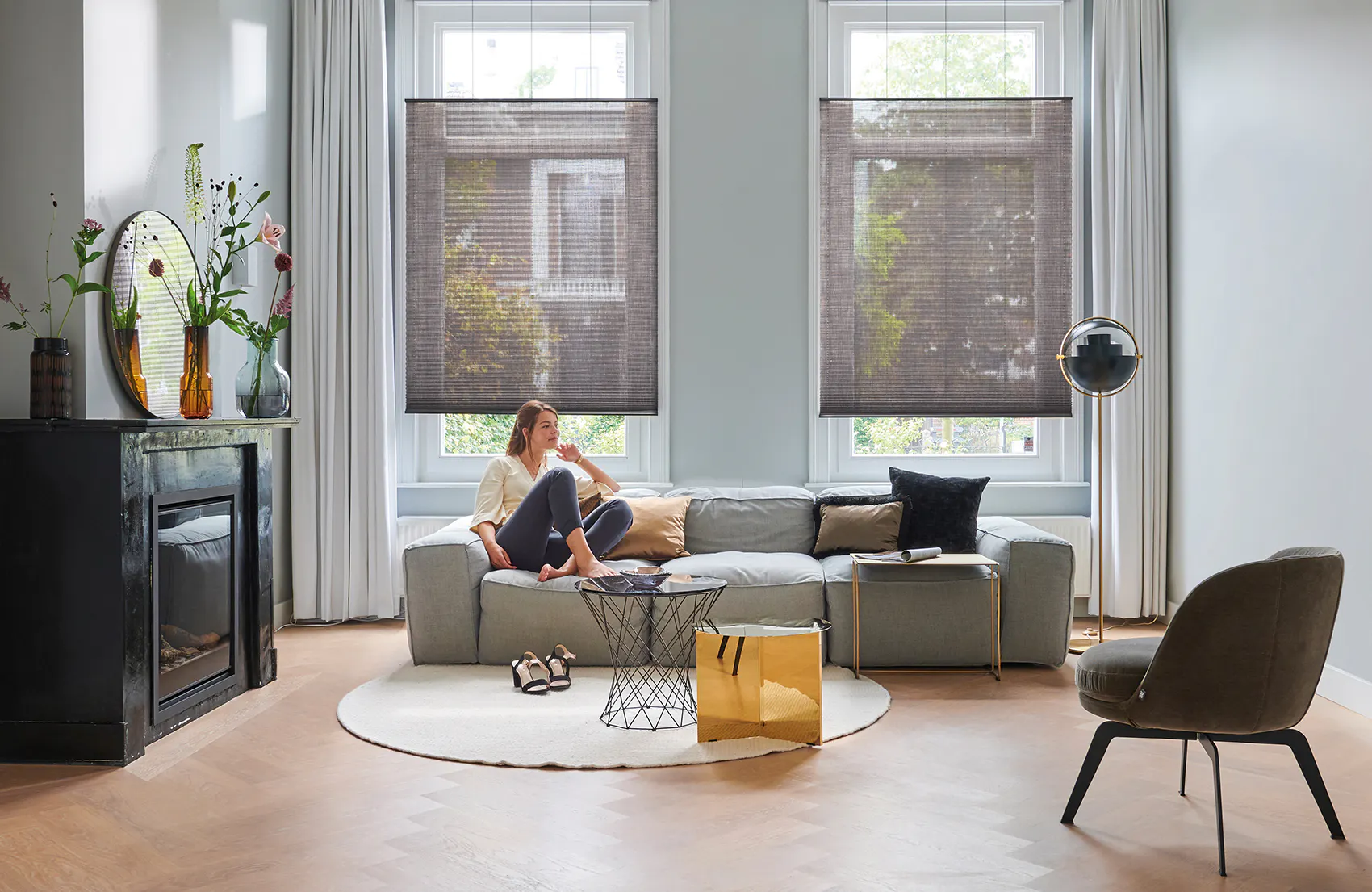 To halvtransparente brune rullegardiner dekker store vinduer mens en kvinne sitter i sofa og leser; lyse sidegardiner, moderne stue med peis, rundt teppe, sofabord og lenestol.