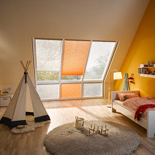 To skråtaksvinduer med delvis nedsenkede persienner slipper inn mykt dagslys i et lyst loftssoverom med oransje vegger, seng, nattbord, lampe og stol.