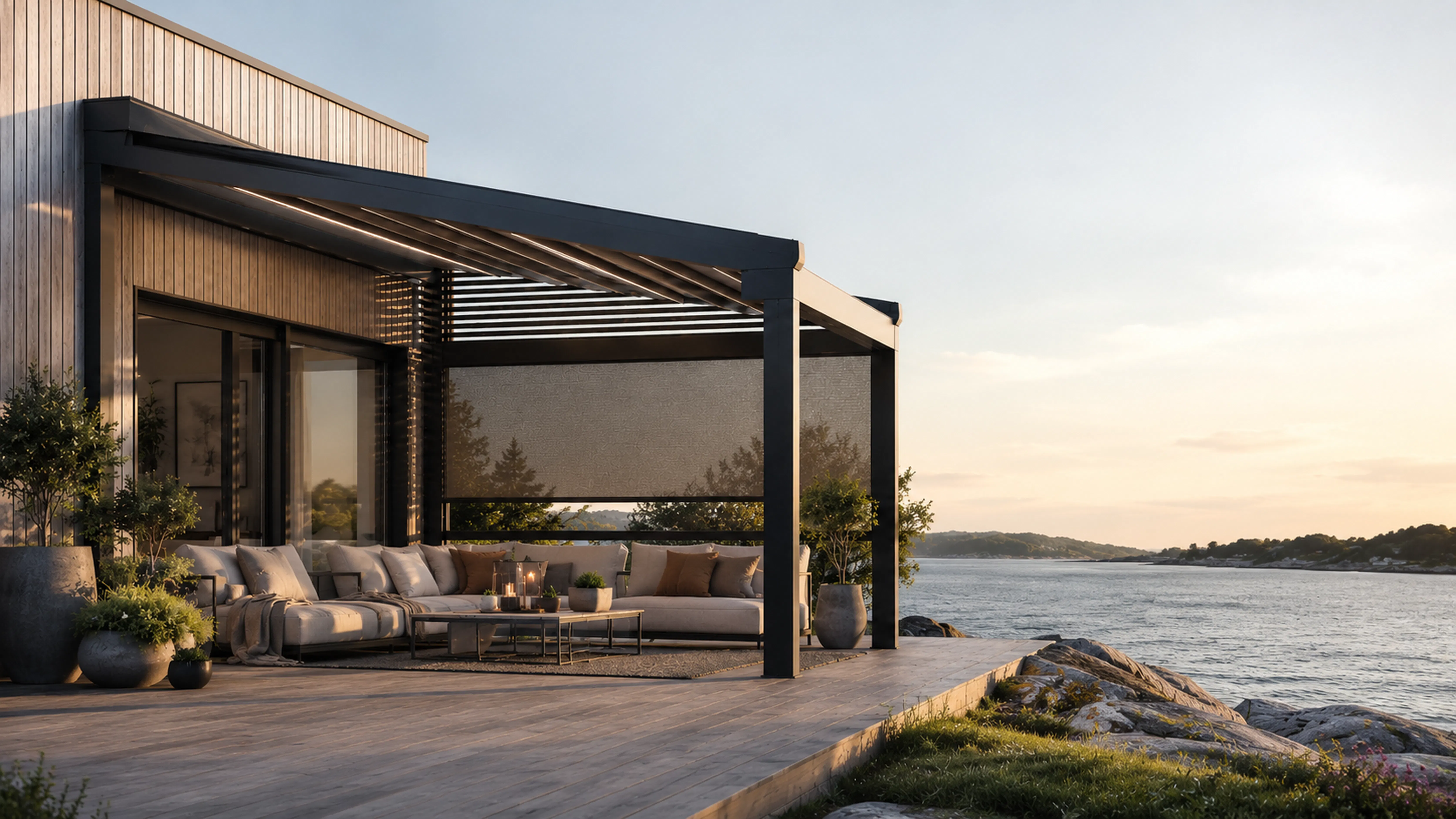 Pergola med nedrullede utendørs screens gir skygge og privatliv over en stor hjørnesofa; moderne terrasse ved kysten med steiner og rolig sjø i solnedgang.