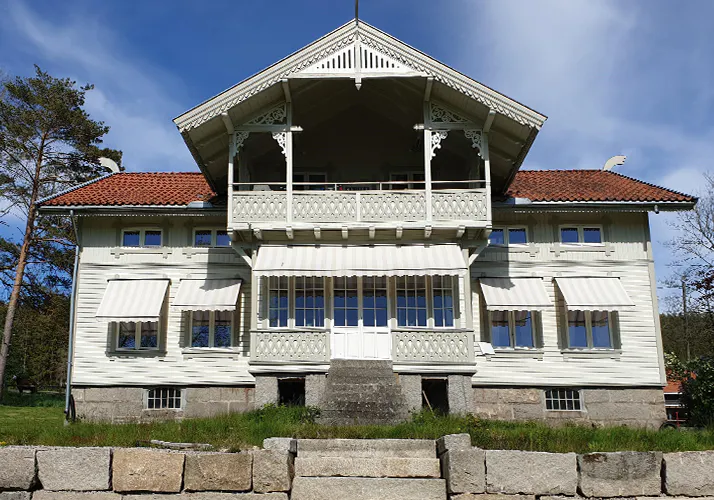 Stripete vindusmarkiser dekker flere vinduer på en stor hvit, dekorativ trevilla; markisene er trukket ut og gir skygge. En utskåret balkong og steintrapp står i forgrunnen under klar, blå himmel.