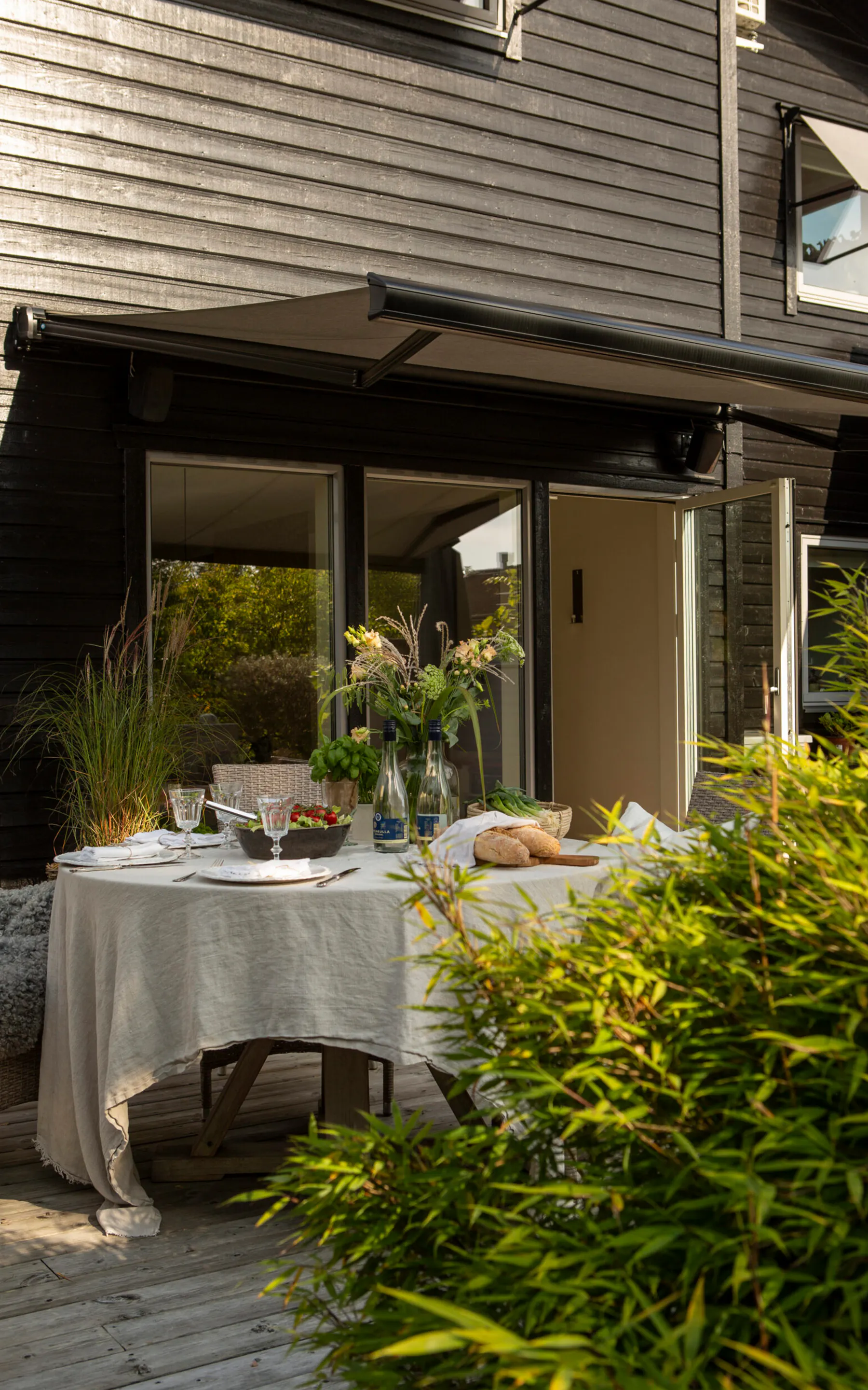 En utstrakt markise skygger et hvitt duket spisebord på treterrassen utenfor et mørkt, panelkledd hus med åpne dører, kurvmøbler og frodige potteplanter.