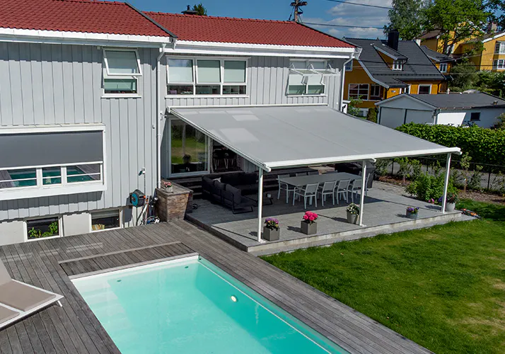 En stor lysegrå markise dekker en terrasse og gir skygge over spisebord og stoler ved et hvitt to-etasjes hus; foran ligger et svømmebasseng og en grønn plen.