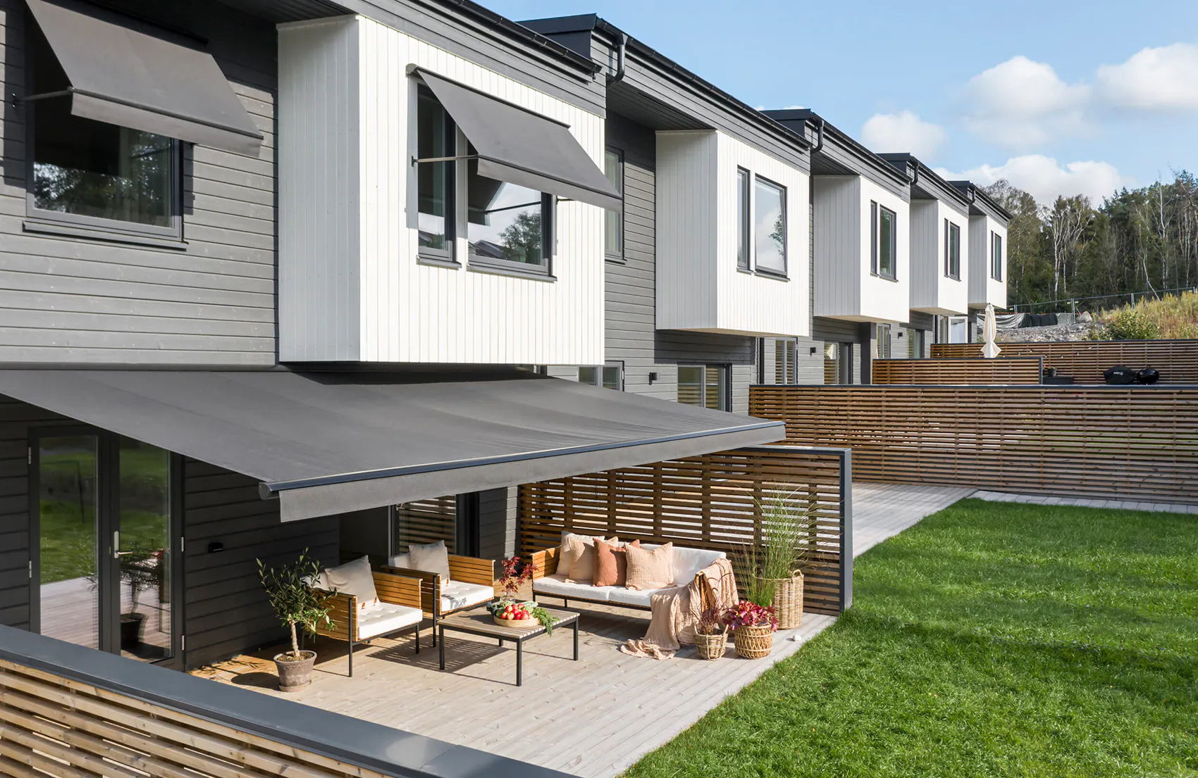 Markiser er trukket ut over terrasse og vinduer, skygger en sittegruppe med sofa og bord ved moderne rekkehus, med grønt gress og trehekk i bakgrunnen.