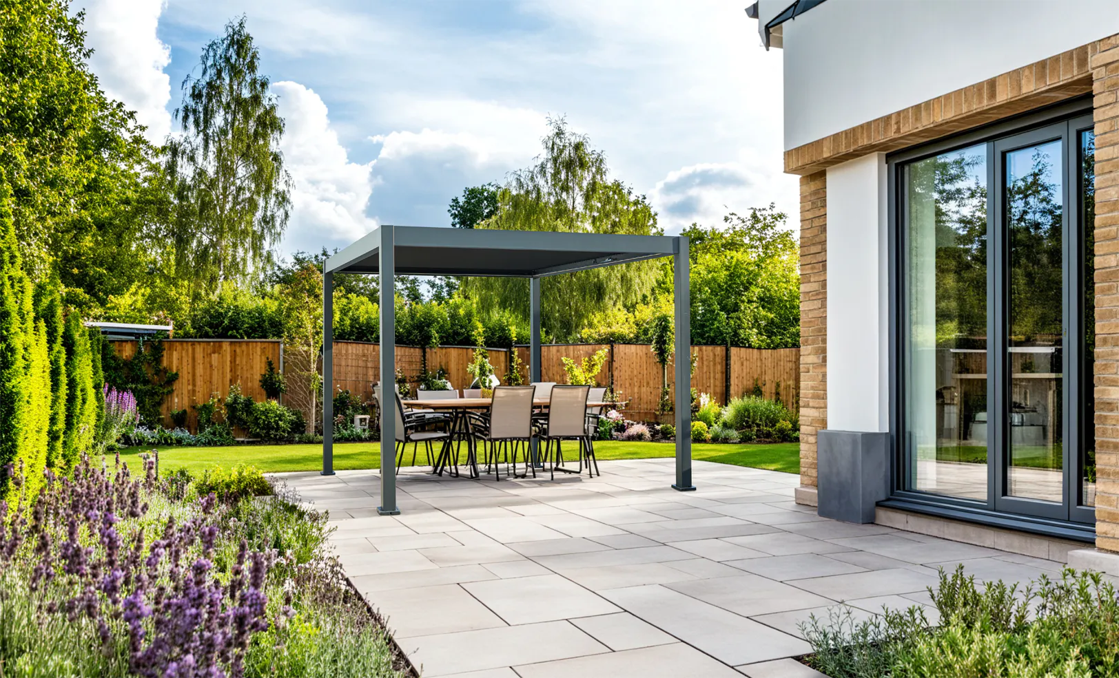 En frittstående pergola gir skygge over et utebord med seks stoler på steinlagt patio ved en moderne husfasade, omgitt av plen, hekk og blomsterbed i en solrik hage.