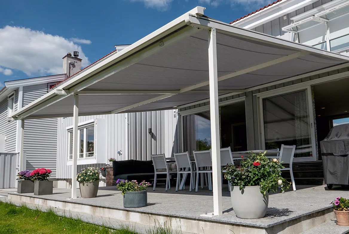 Terrassetak/markise strekker seg over en betongterrasse, skygger et spisebord med stoler; hvitt trehus i bakgrunnen og flere blomster i potter langs terrassekanten.