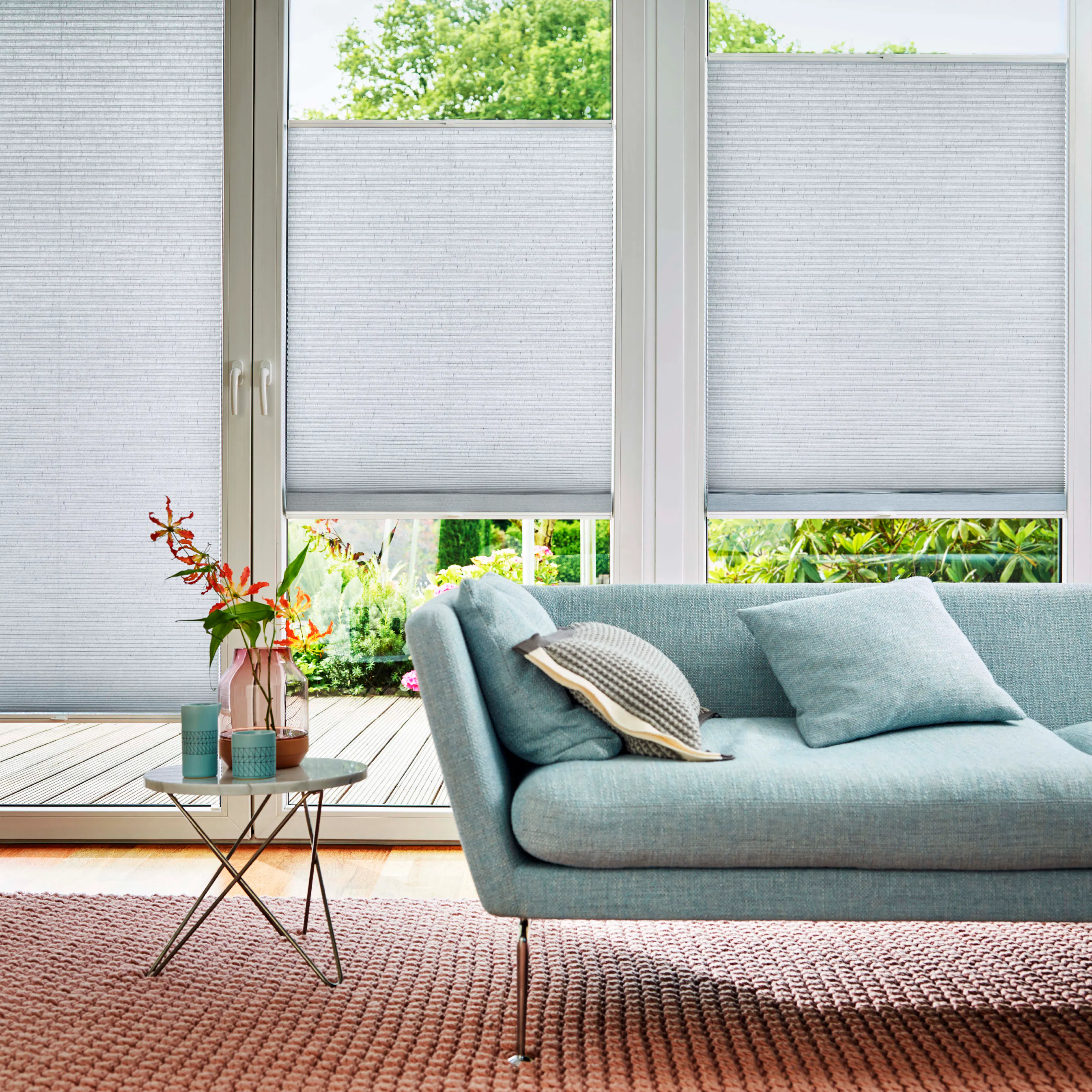 Hvite plisségardiner er delvis senket foran store vinduer i en lys stue; lyseblå sofa med puter, lite sidebord med vase og kopper, rosa teppe og utsikt mot grønn hage.