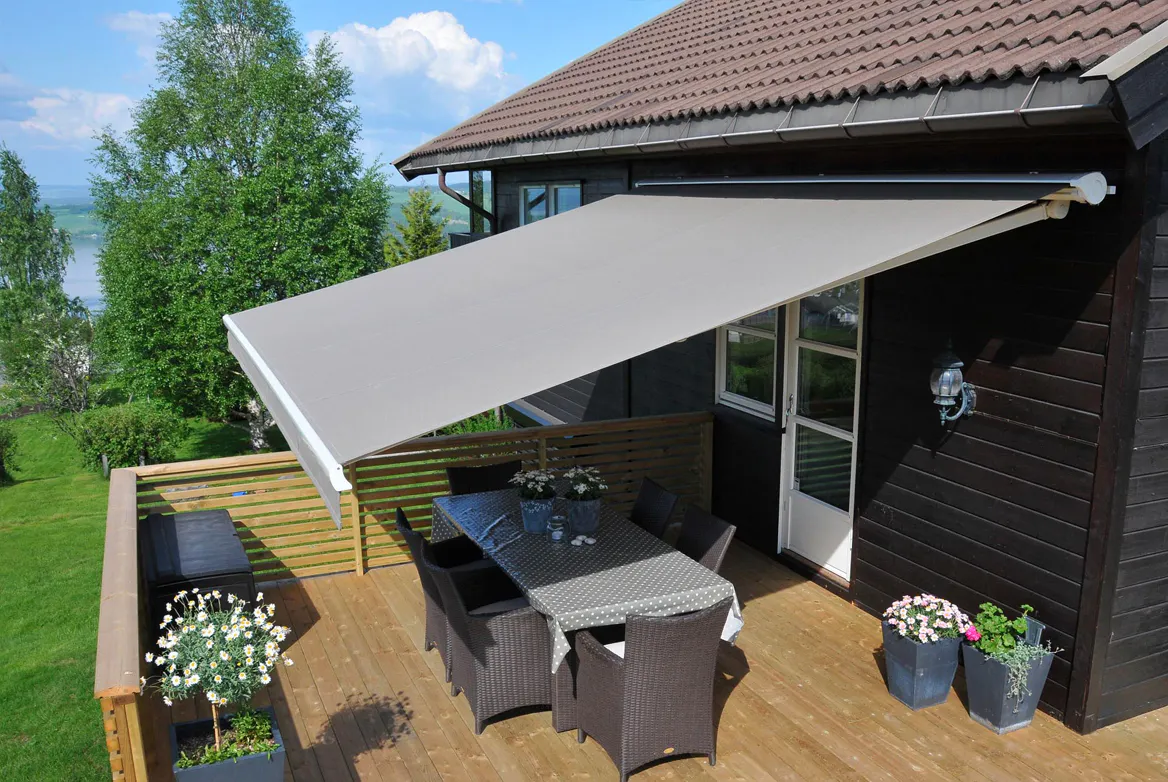 En grå kassettmarkise dekker og gir skygge over et utebord med seks stoler på en treterrasse festet til et mørkt trehus, omgitt av blomsterkrukker, gress og trær.